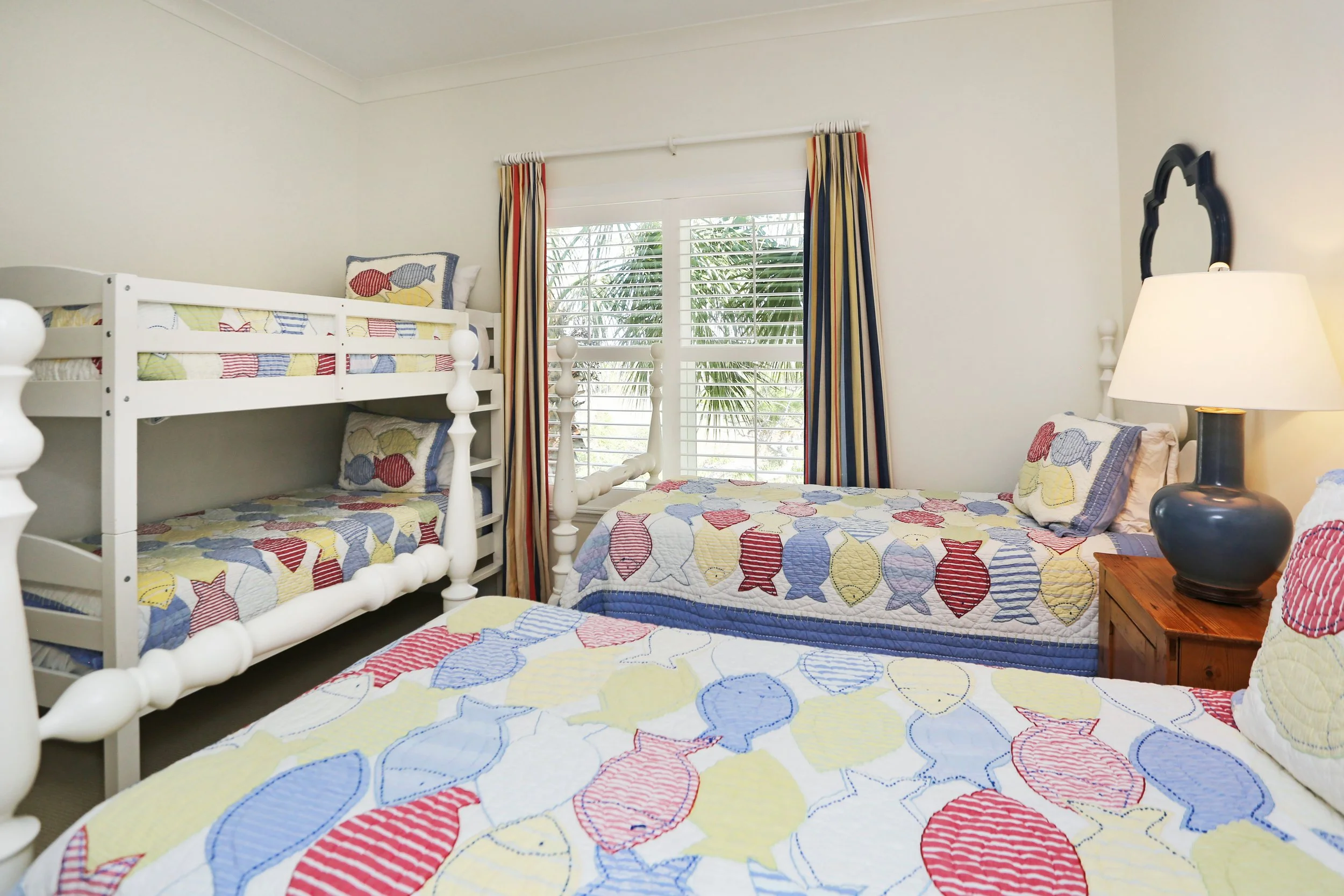 A bedroom with two beds, a bunk bed and a single bed, featuring colorful fish-patterned quilts and pillows. There is a window with curtains and a nightstand with a lamp between the beds.