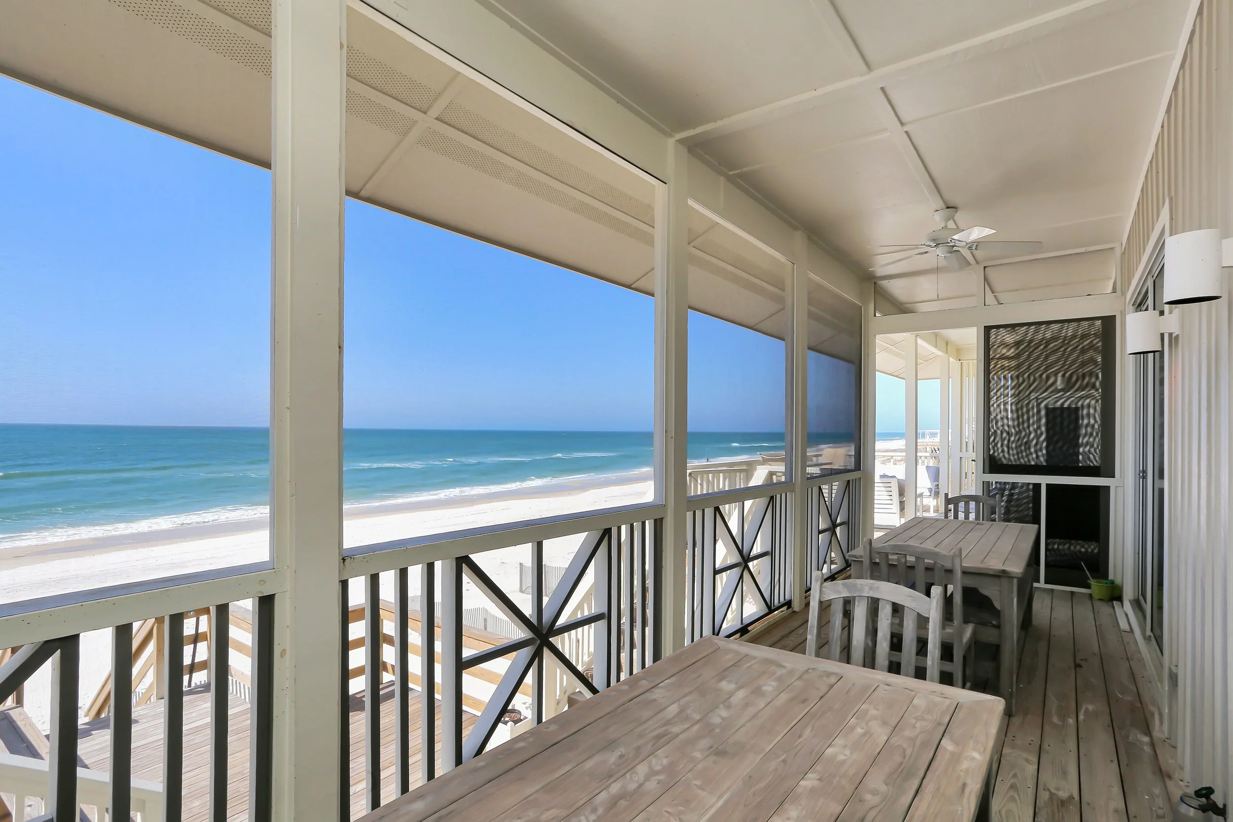 Ocean view from a beach balcony with wooden tables and chairs, overlooking a sandy beach and blue water under a clear sky.