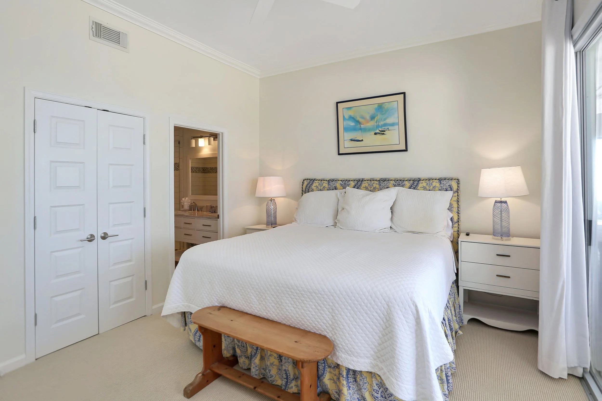 A bedroom with a bed, two nightstands with lamps, a framed picture of sailboats above the bed, a wooden bench at the foot of the bed, a closet with double doors, and a view of a bathroom through an open door.