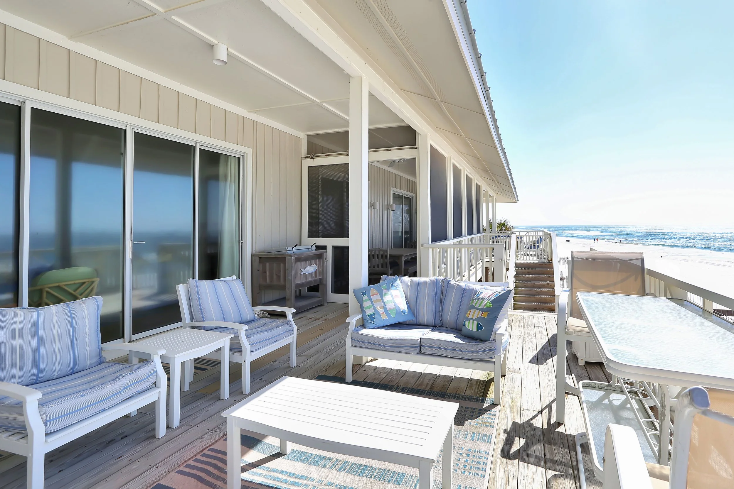Beachside balcony with outdoor furniture including cushioned chairs, a loveseat, a table, and a dining table with chairs, overlooking ocean waves and sandy shore.