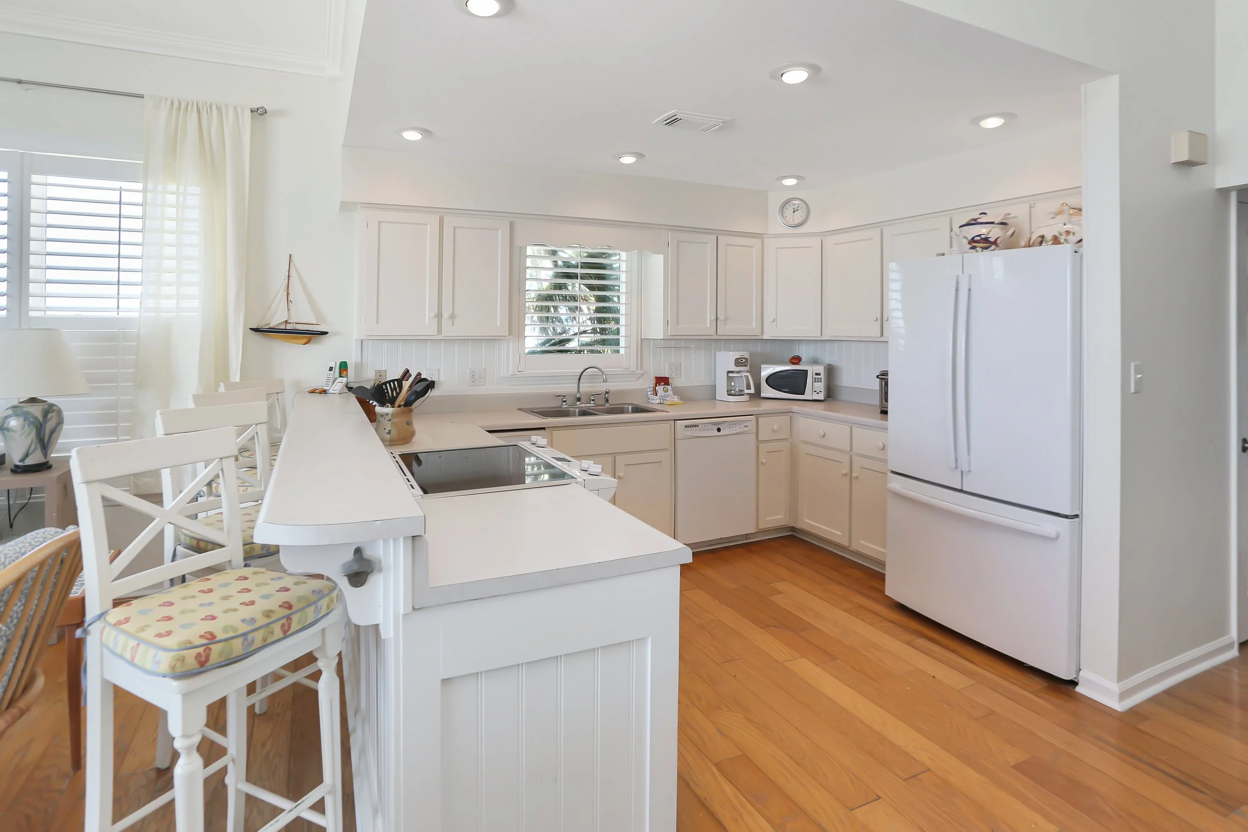 Bright white kitchen with wooden flooring, white cabinets, a large refrigerator, microwave, and small appliances on the counter. There are island chairs with patterned cushions and decorative nautical accents.