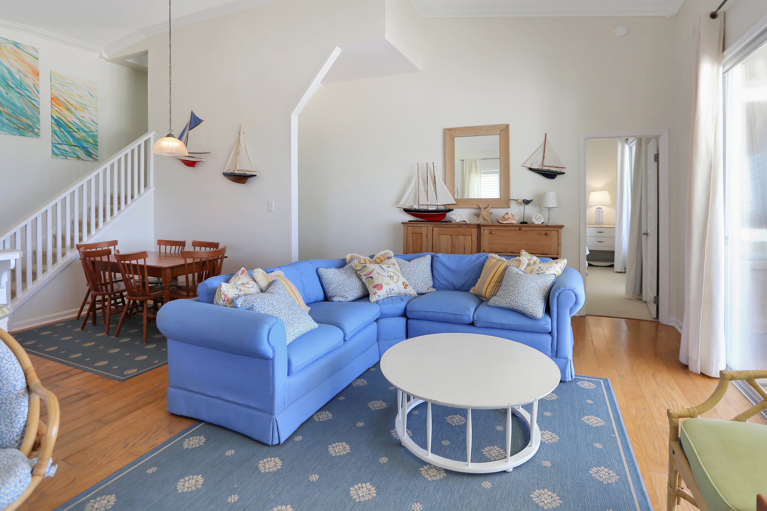 Living room with a blue sectional sofa, decorative pillows, a white round coffee table, a wooden sideboard with model sailboats, a mirror, framed nautical paintings, and a staircase with artwork on the wall.