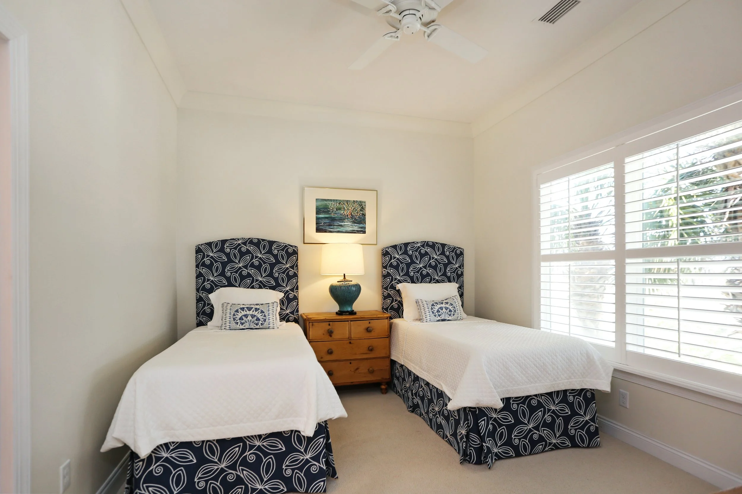 A bedroom with two beds featuring matching dark blue and white patterned headboards, a wooden nightstand with a lamp between the beds, a framed painting above the lamp, and large windows with white blinds.