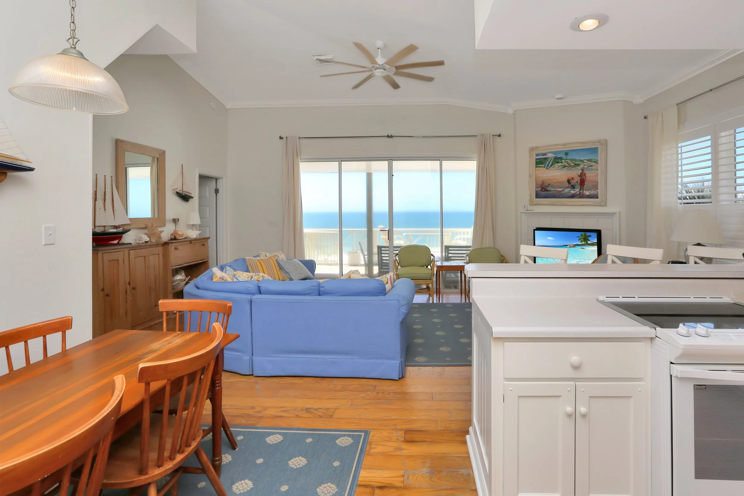 Open-concept living room and kitchen with sliding glass door leading to a balcony with ocean view. Includes a blue sofa, wooden dining table, and coastal decor.