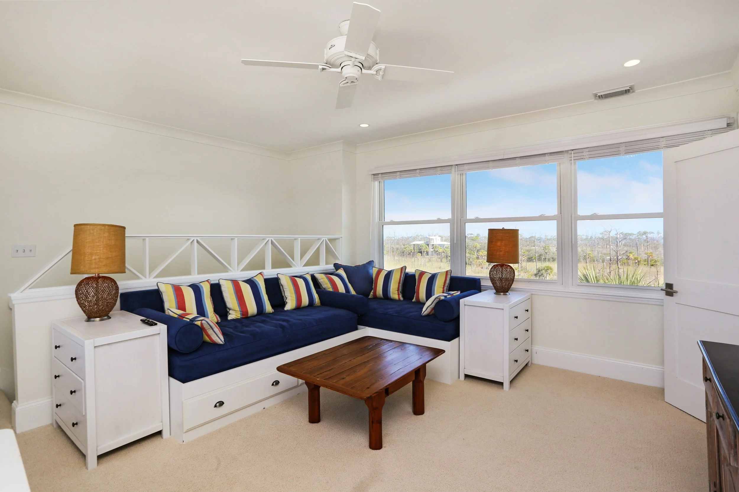 Living room with a navy blue sectional sofa, colorful striped pillows, white side tables with lamps, a small wooden coffee table, large windows showing an outdoor view, and a ceiling fan.
