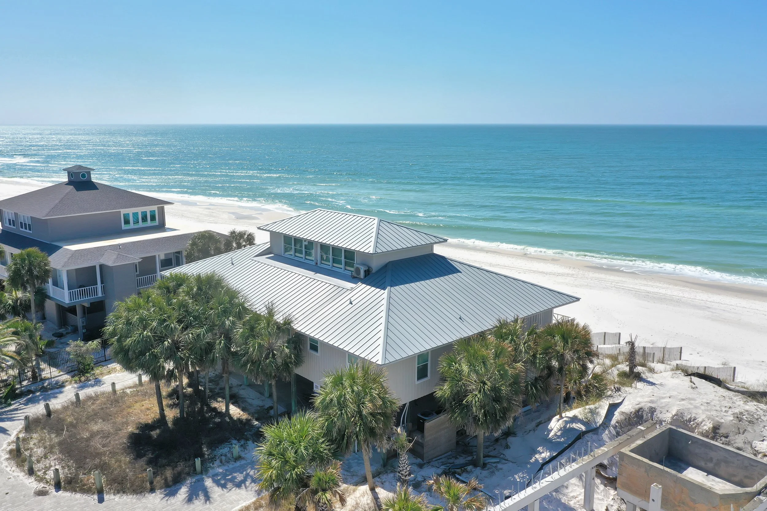 Beachfront houses with palm trees along white sandy beach and ocean waves in background under clear blue sky.