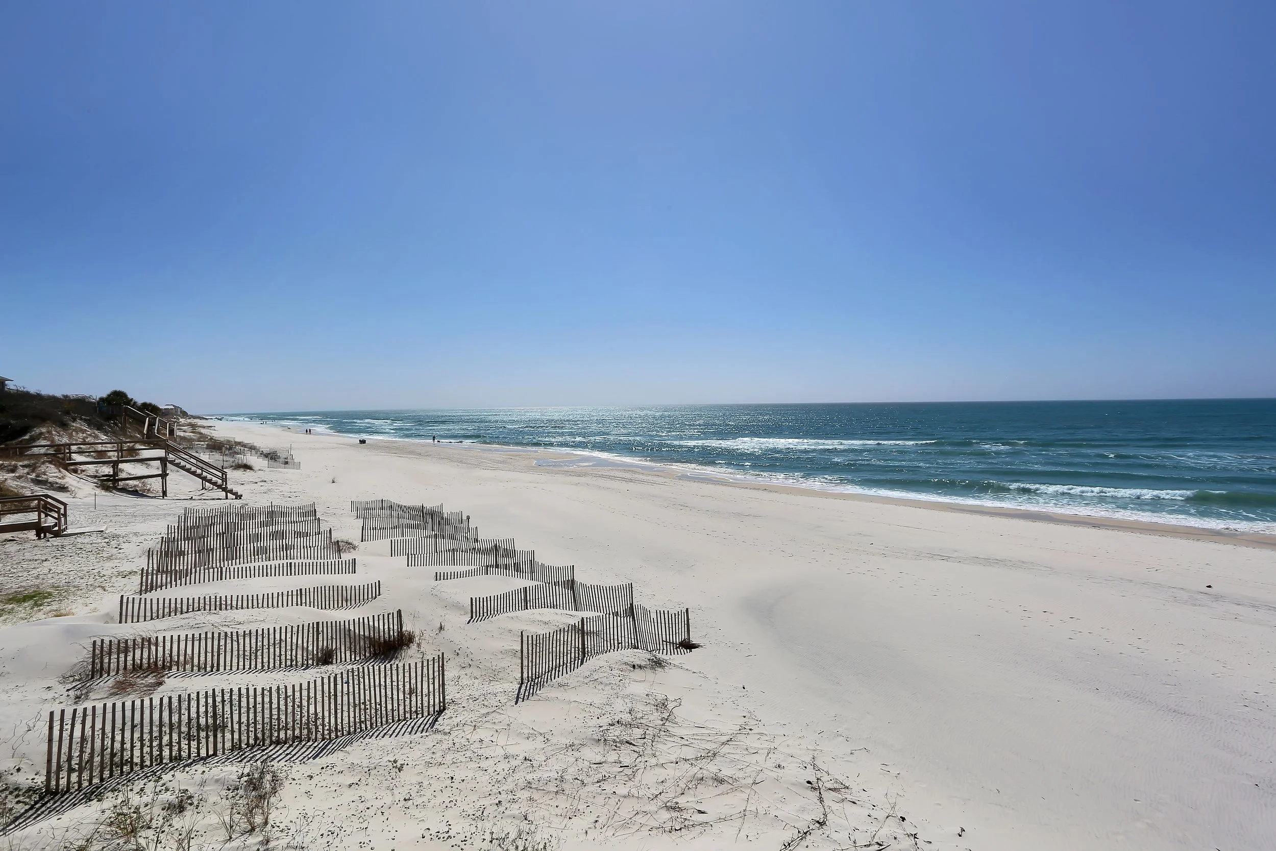 Clean, sandy beach with wooden fences, clear blue sky, and calm ocean waves.