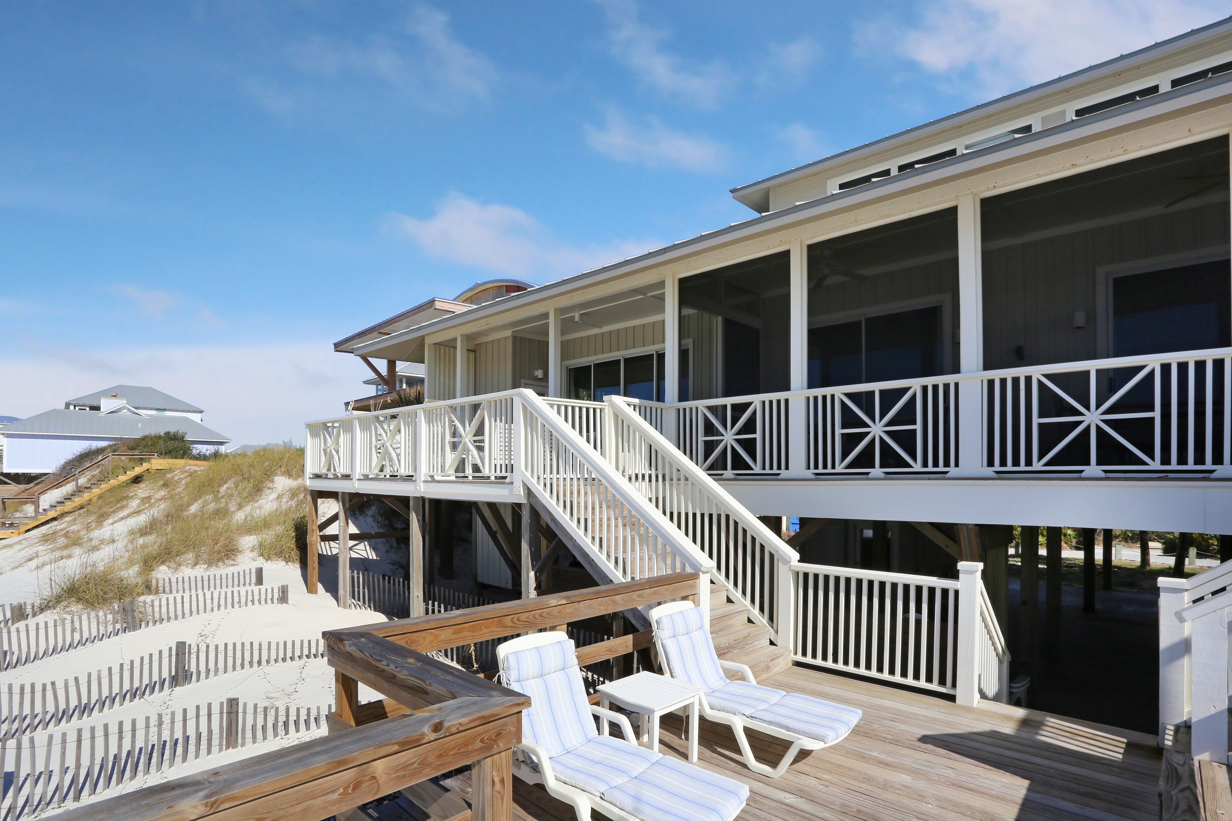 Beach house with a deck and stairs leading to sand, outdoor lounge chairs, and a wooden fence, on a sunny day with clear blue sky.
