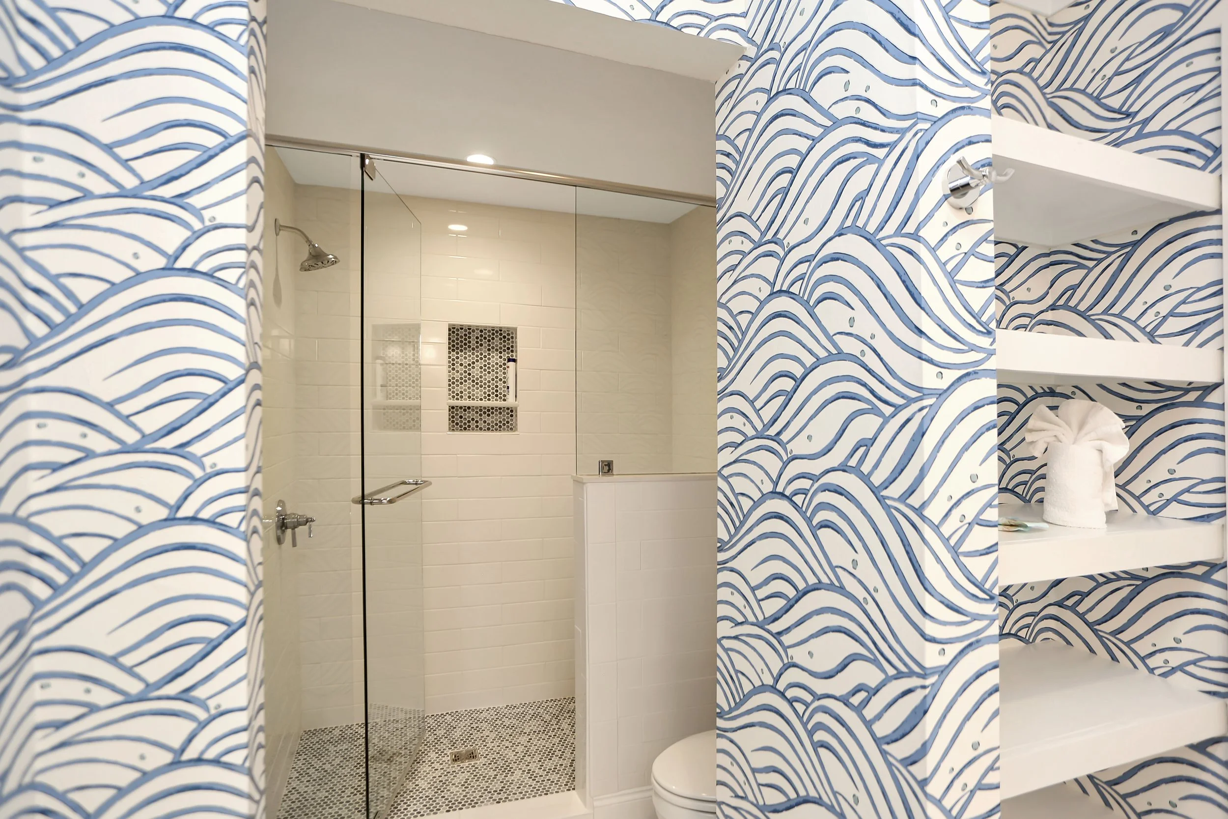 A bathroom with a glass shower enclosure, white tiled walls, and a showerhead. The walls outside the shower feature blue and white wave-patterned wallpaper. There are white shelves with a rolled towel and some toiletries.