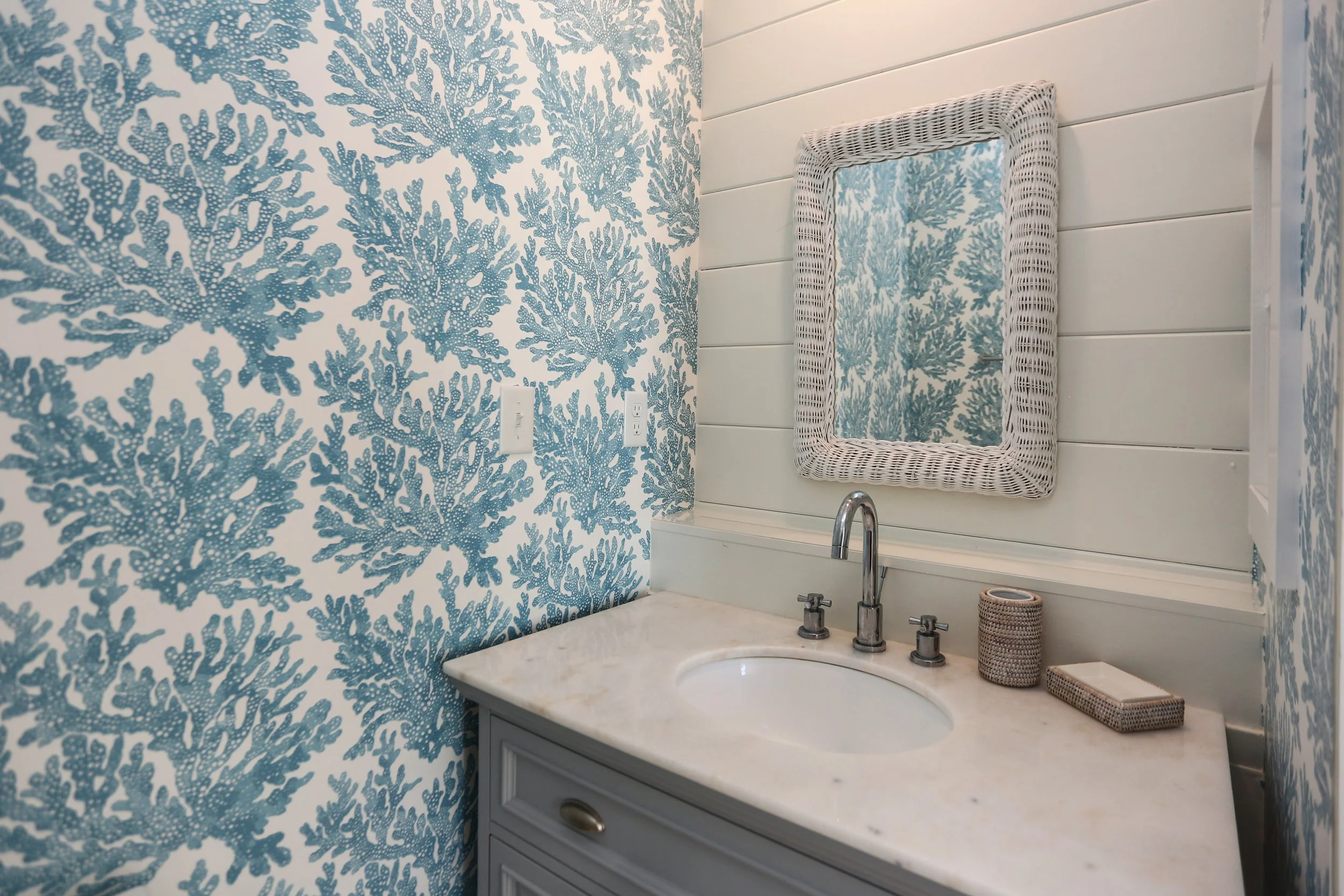 Bathroom with blue coral-patterned wallpaper, white sink with marble top, wicker framed mirror, and small wicker accessories on the counter.