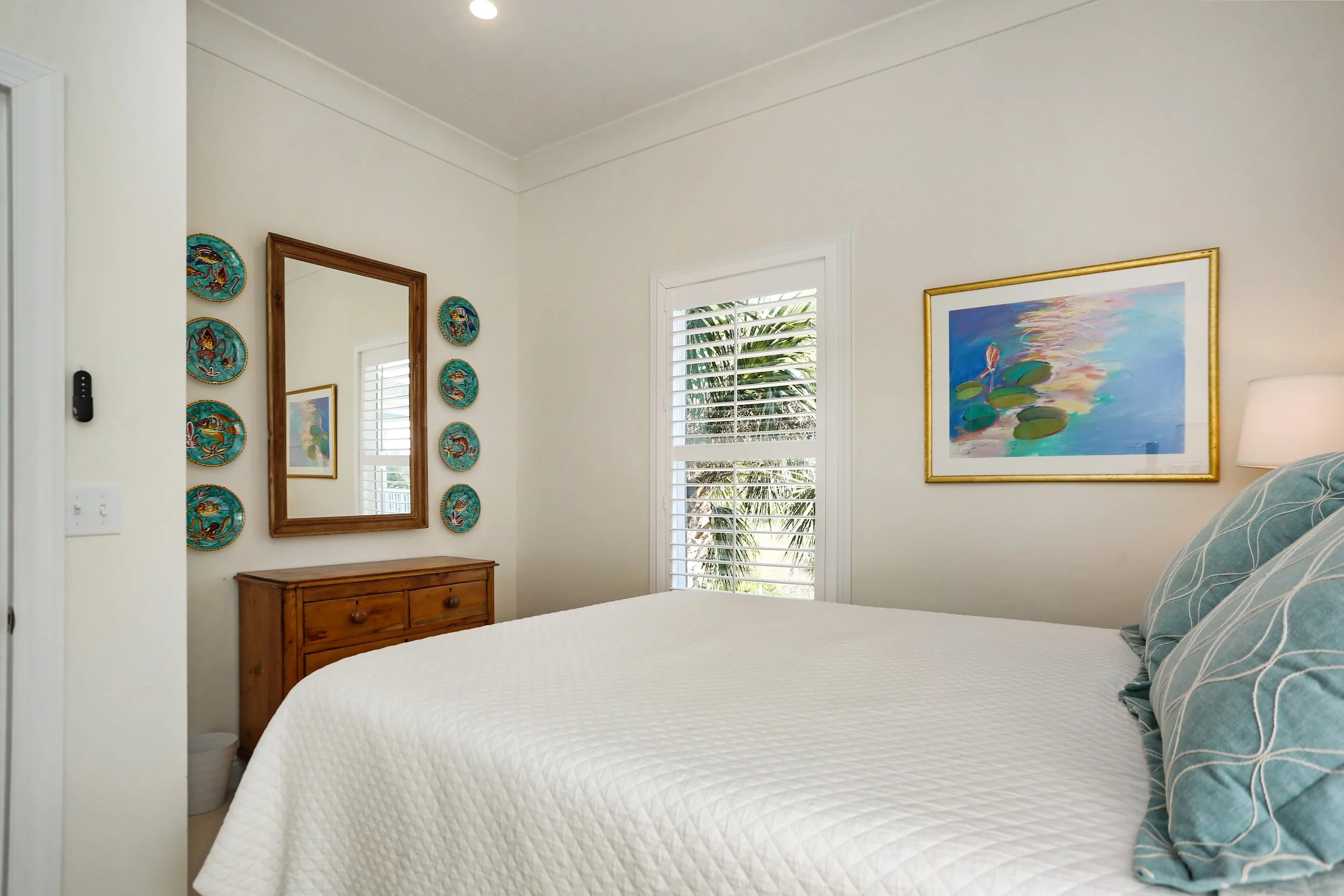 A bedroom with a white quilted bed, blue patterned pillows, a wooden dresser, a framed picture of water lilies, a mirror surrounded by decorative plates, and a window with white shutters showing palm trees outside.