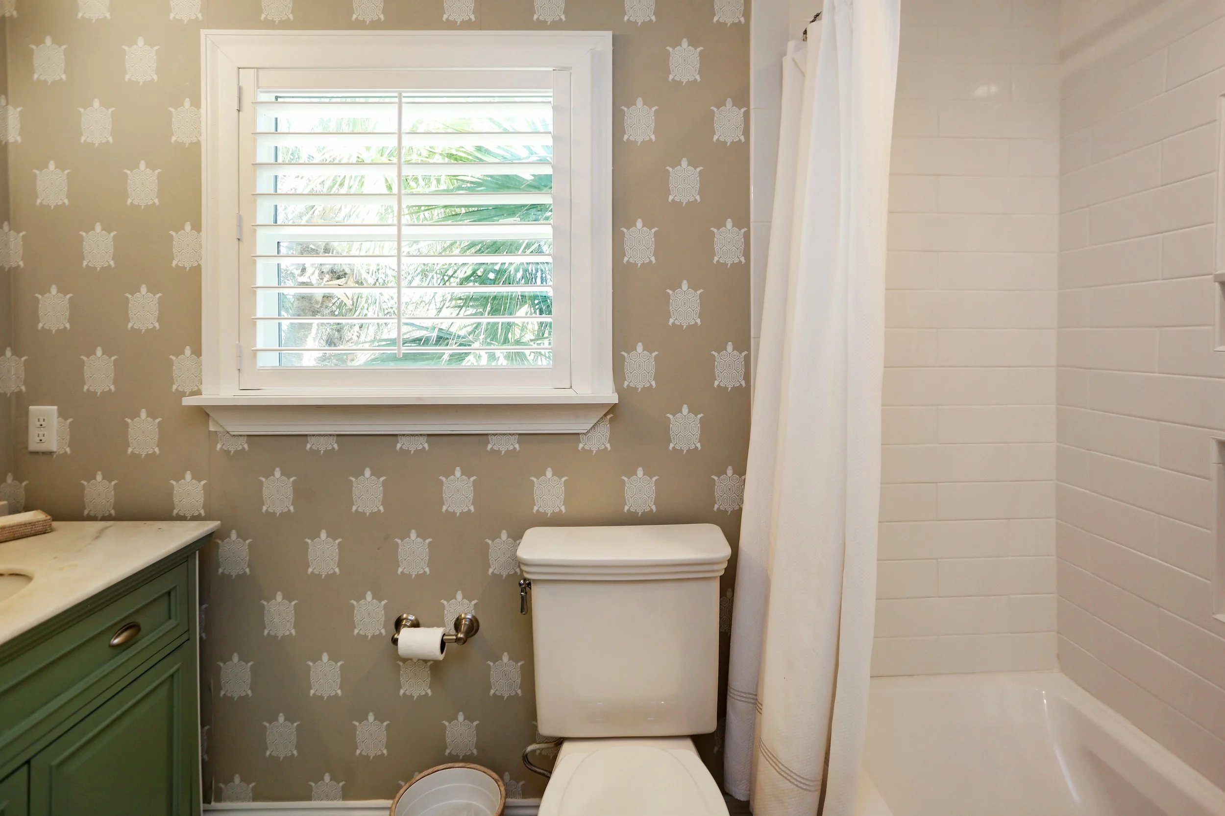 A small bathroom with a window, patterned wallpaper, a toilet, a green vanity, and a shower with white tiles.
