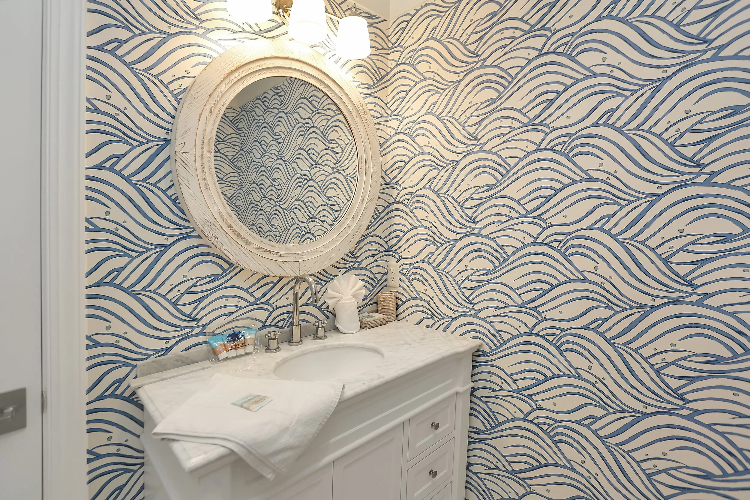 Bathroom sink with a white vanity, a marble countertop, a round mirror with a white frame, and blue wave-patterned wallpaper.