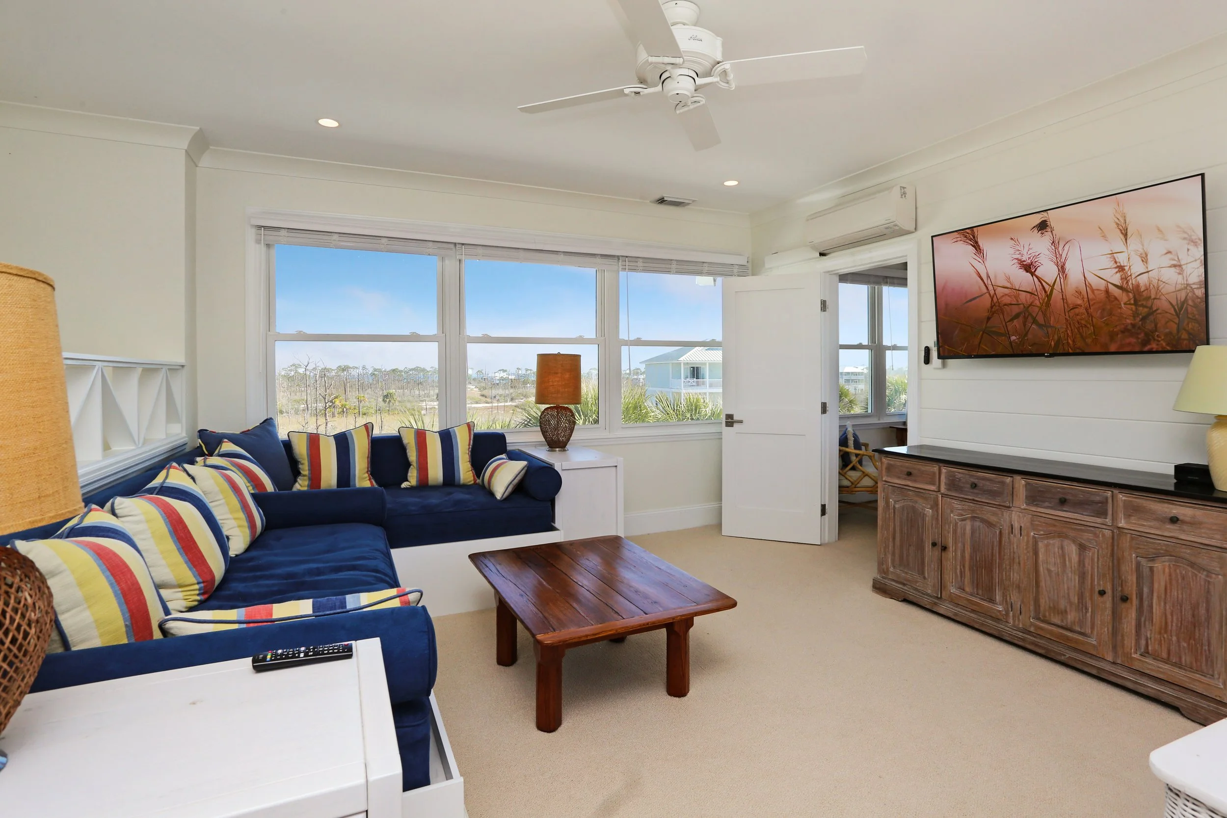 Coastal living room with beige carpet, white walls, large windows showing the outdoors, navy sectional sofa with striped yellow, red, and blue cushions, wooden coffee table, wooden sideboard, wall-mounted flat-screen TV, and a ceiling fan.