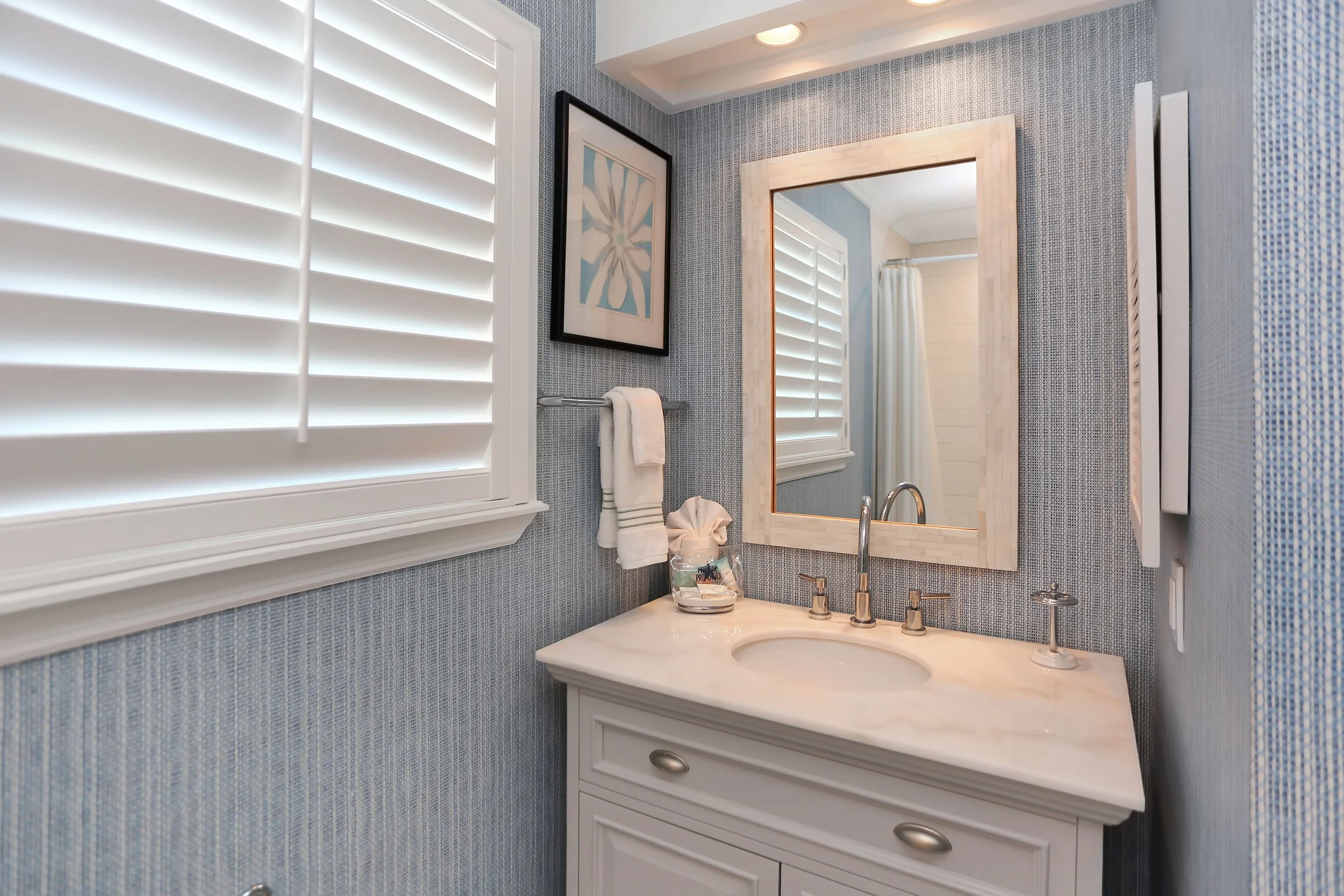 Bathroom vanity with a white countertop, sink, mirror, and a small towel rack with towel, window with white shutters, and framed artwork on a textured blue wall.
