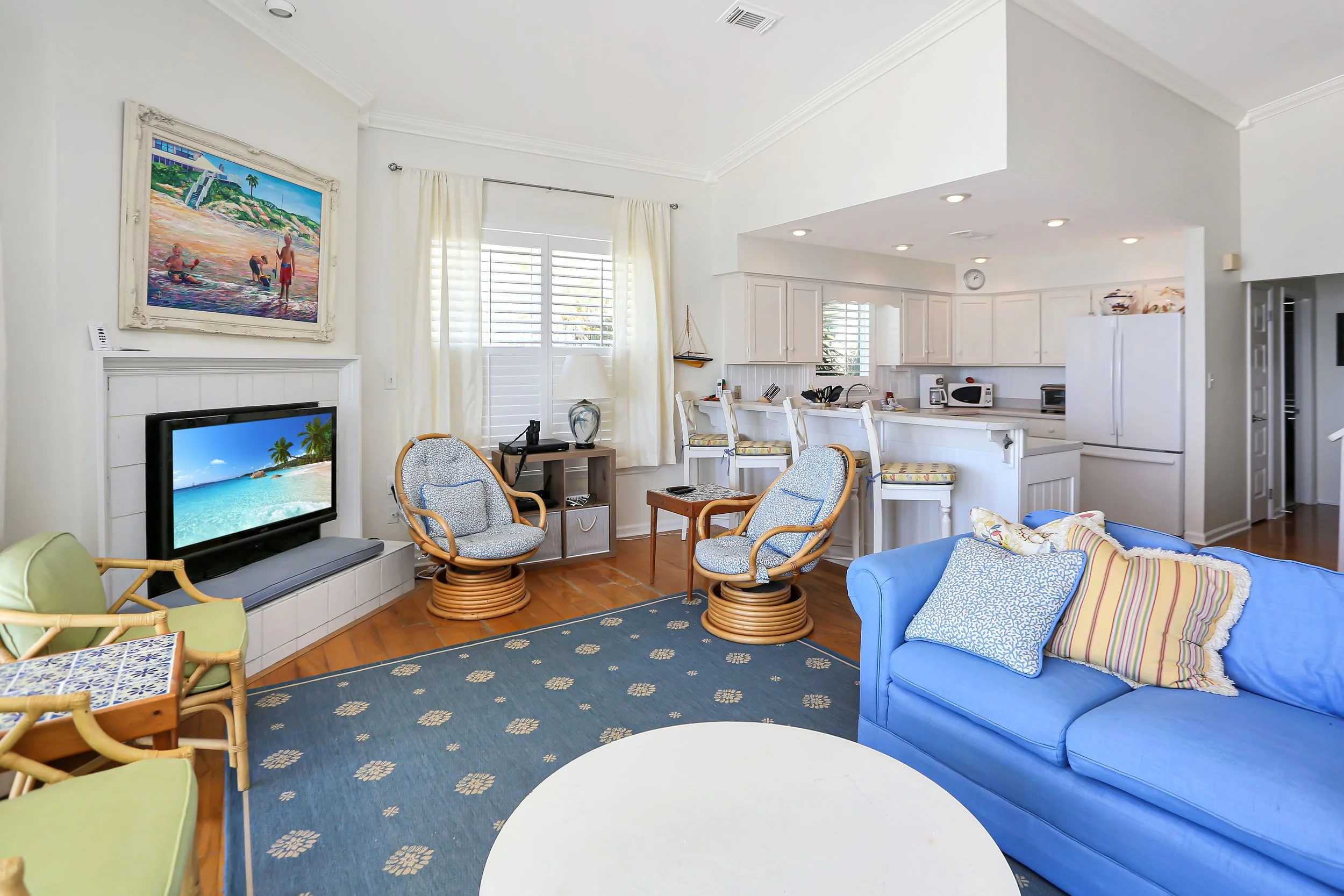 Living room with blue sofa, patterned pillows, wooden chairs, a television showing a beach scene, and a view into the kitchen with white cabinets and stainless steel appliances.