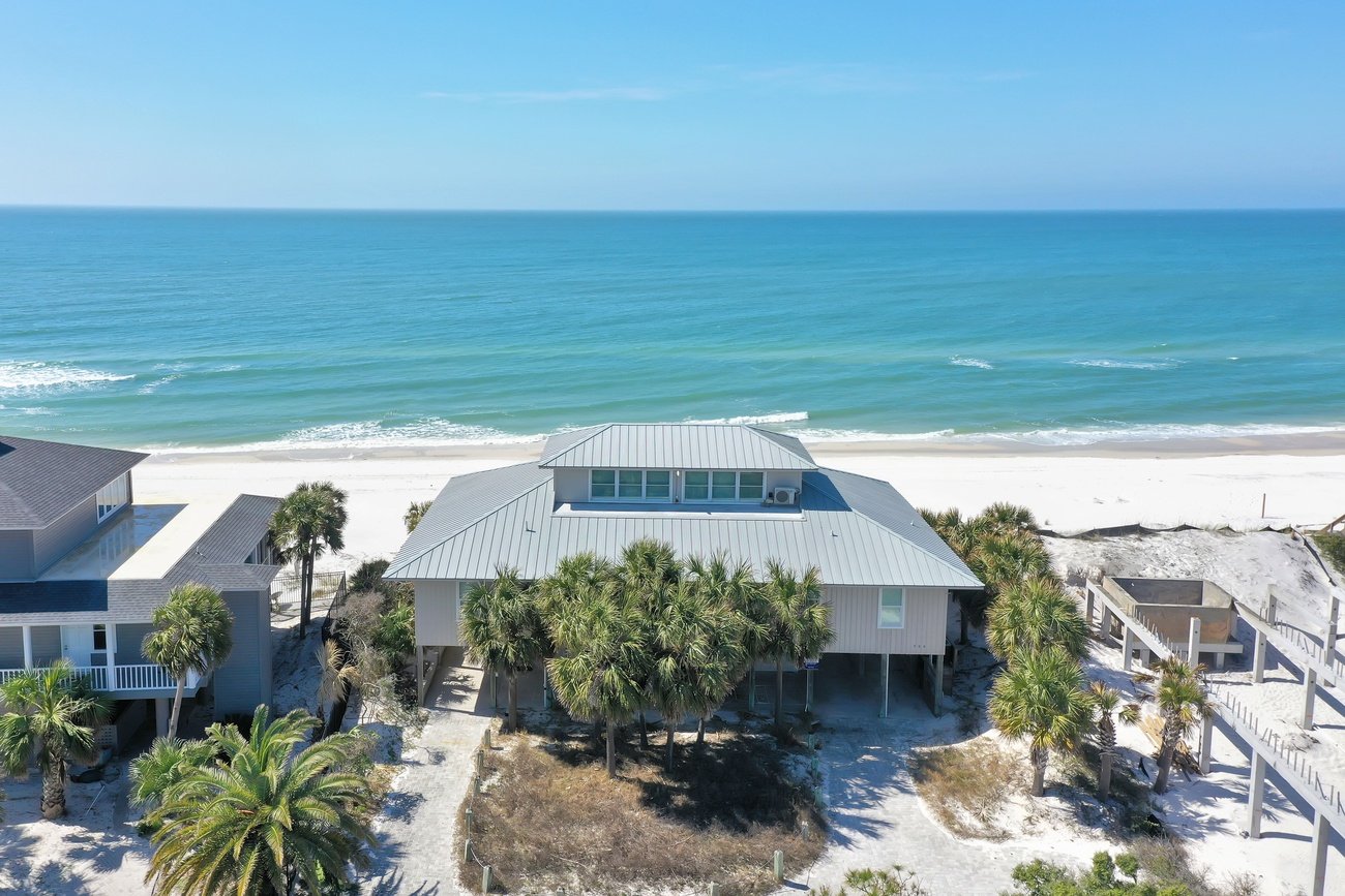 An aerial view of a beach house with a metal roof, surrounded by palm trees, located on a white sandy beach with the ocean in the background.