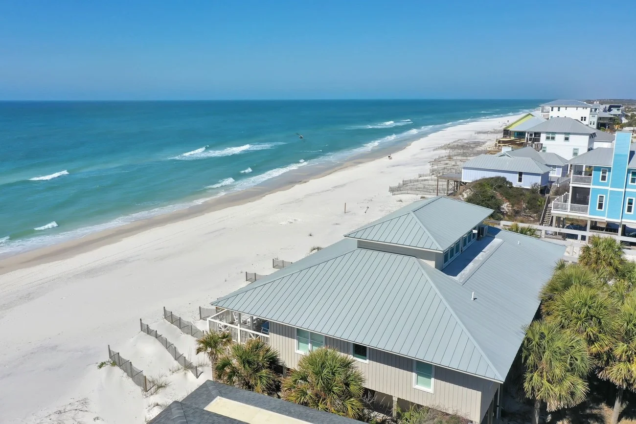 Aerial view of a beach with white sand, ocean waves, and several coastal houses with metal roofs, some palm trees in front.