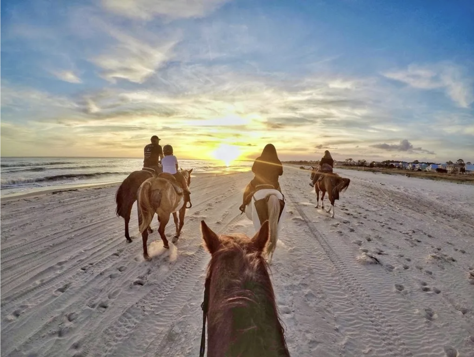 People riding horses on a sandy beach during sunset, with the ocean on one side and a row of small houses in the distance.