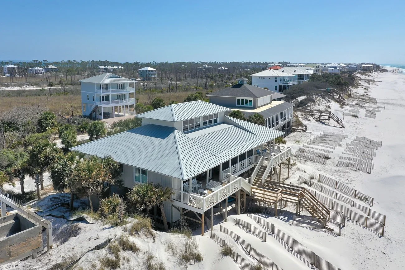 Aerial view of beach houses on a sandy shoreline with wooden stairs and fences, and clear blue sky.