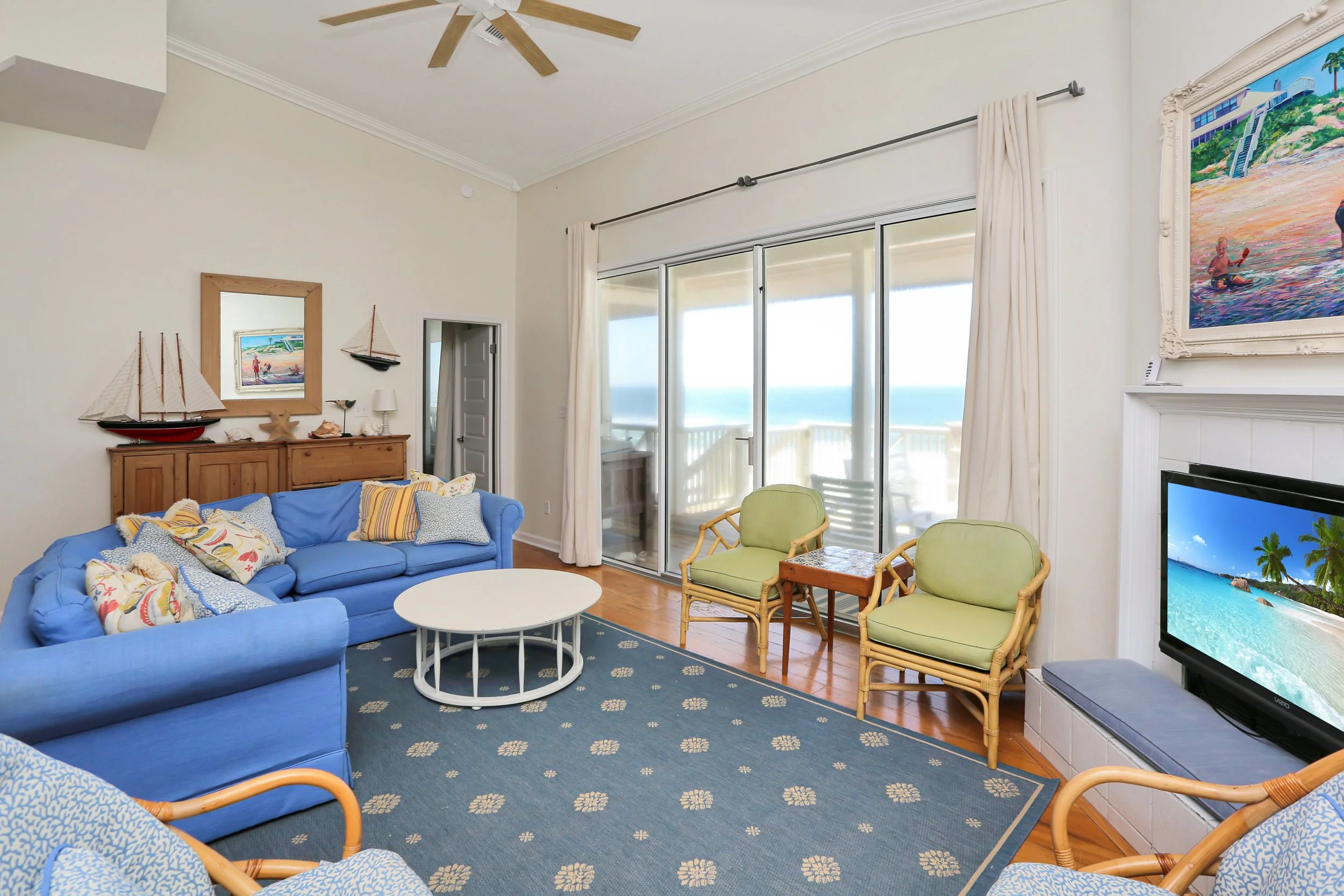 Living room with blue sofa, wicker chairs, nautical decor, sliding glass doors leading to balcony with ocean view, television, and ocean-themed artwork.