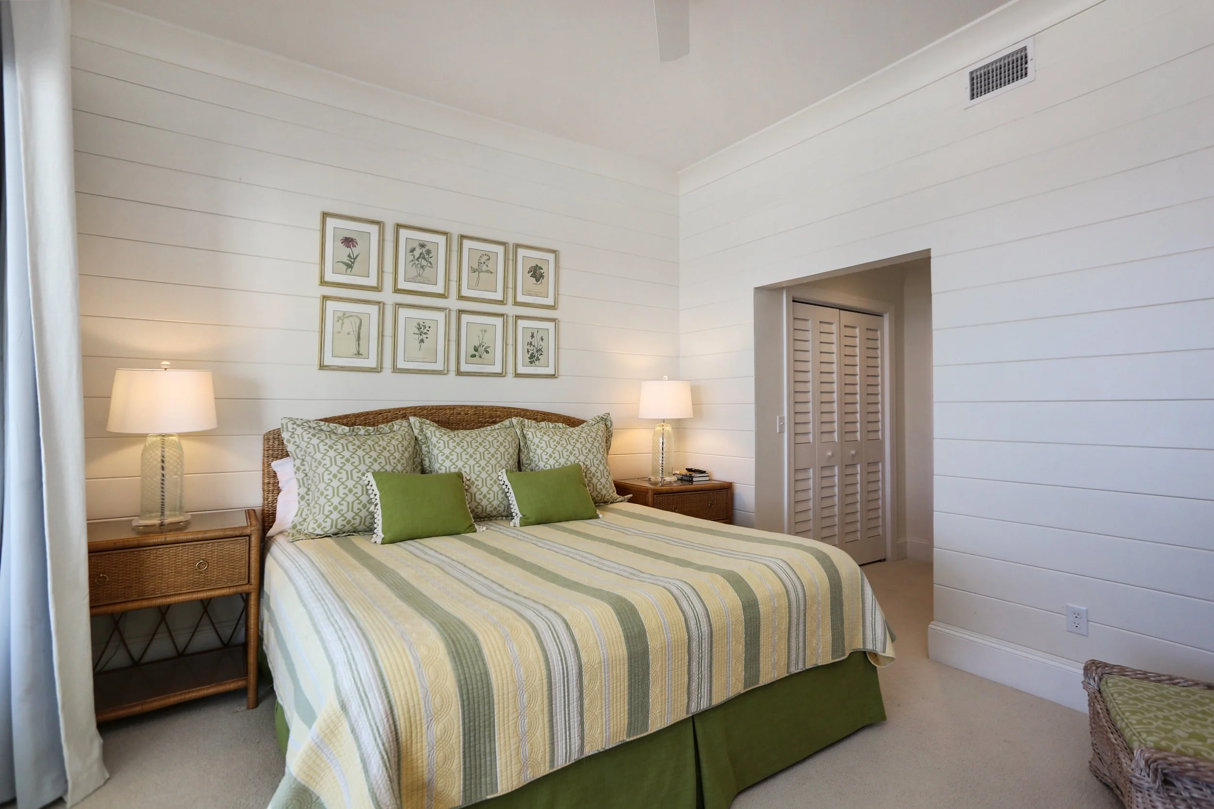 A bedroom with a bed that has green and beige striped bedding, floral pillows, and a wicker headboard. There are two matching bedside tables with lamps, and framed botanical prints on the white-washed wall.
