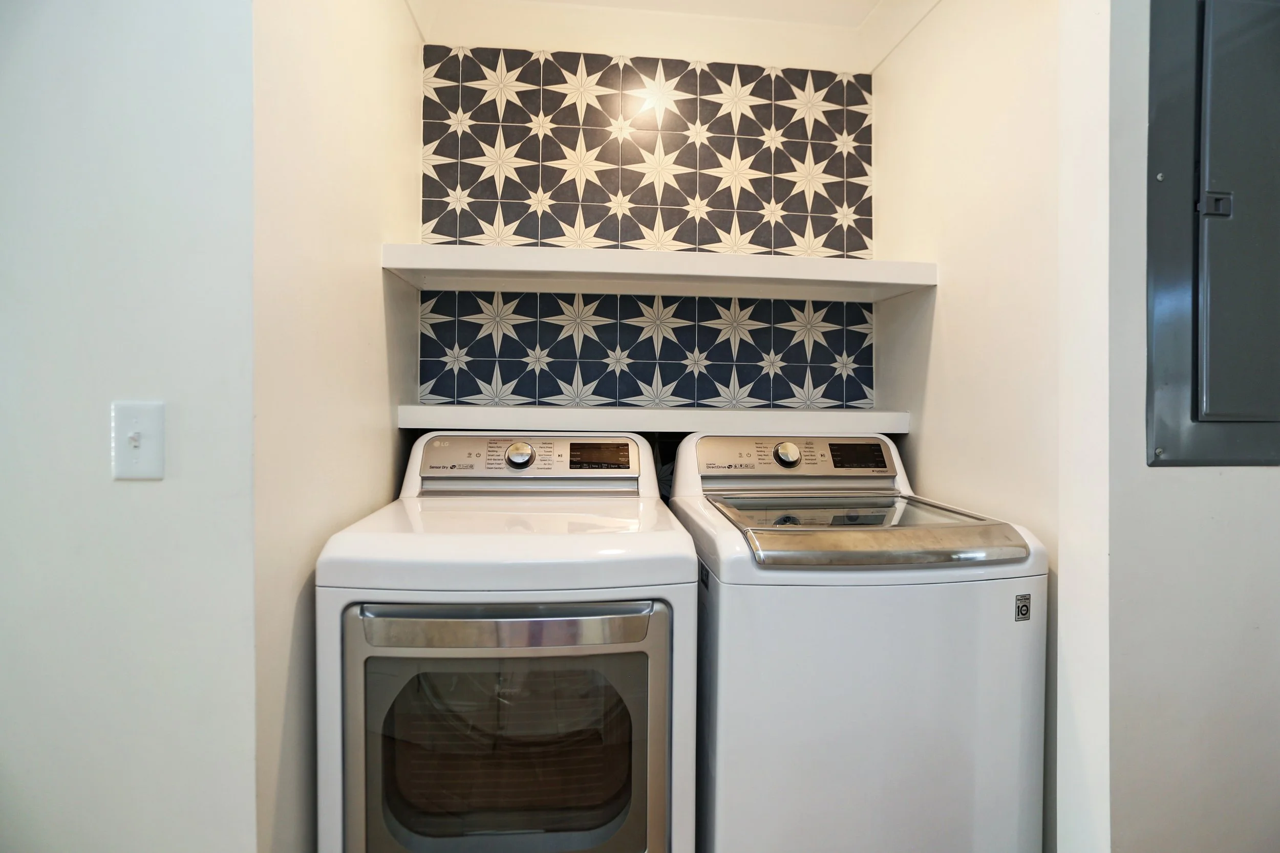 A laundry room with a washer and dryer side by side beneath decorative black and white starburst patterned wall tiles, with two white shelves above.