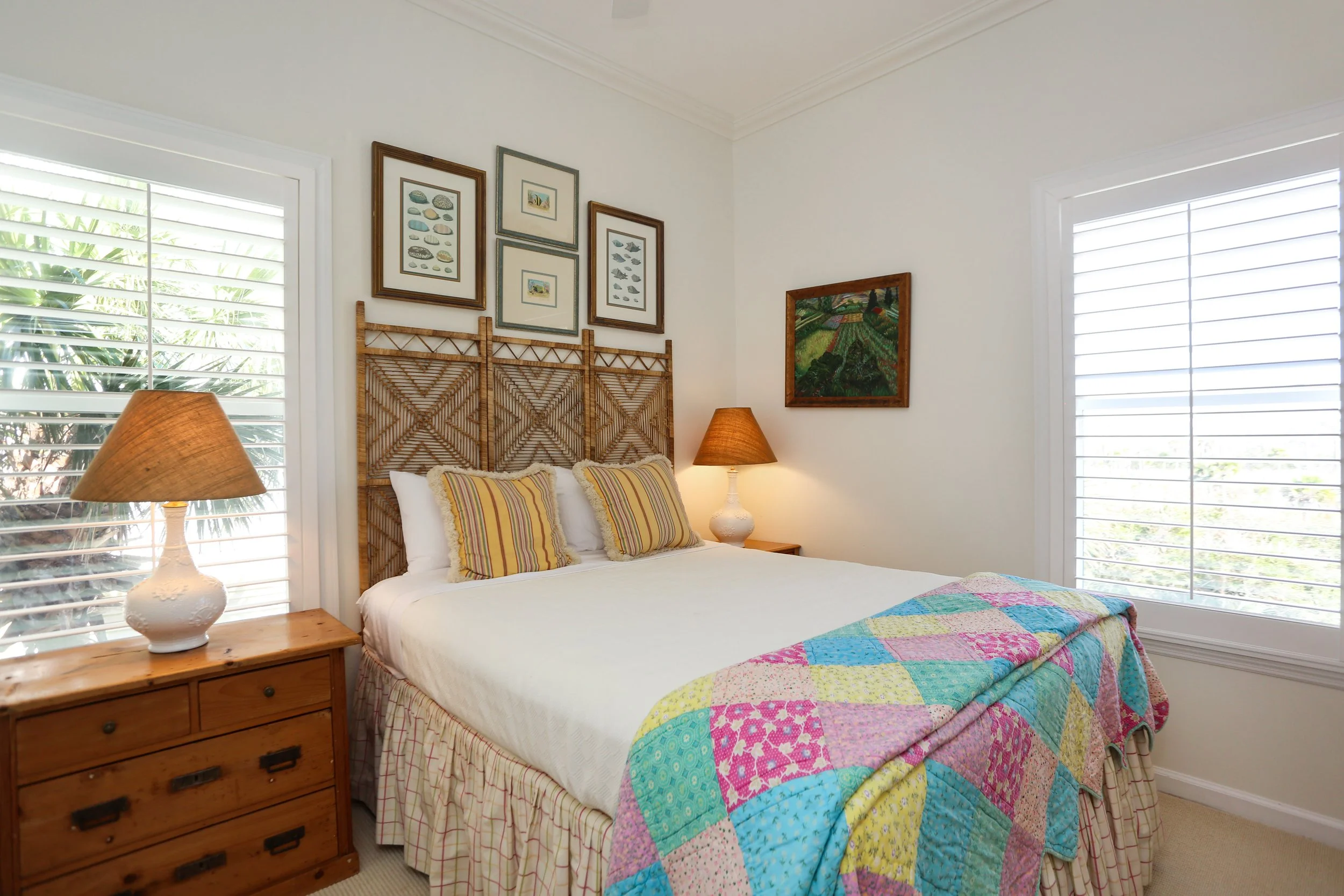 A cozy bedroom with a bed covered with a colorful patchwork quilt. There are three pillows with striped pillowcases on the bed. Two wooden nightstands with lamps sit beside the bed, and framed artwork decorates the white walls. Large windows with whi