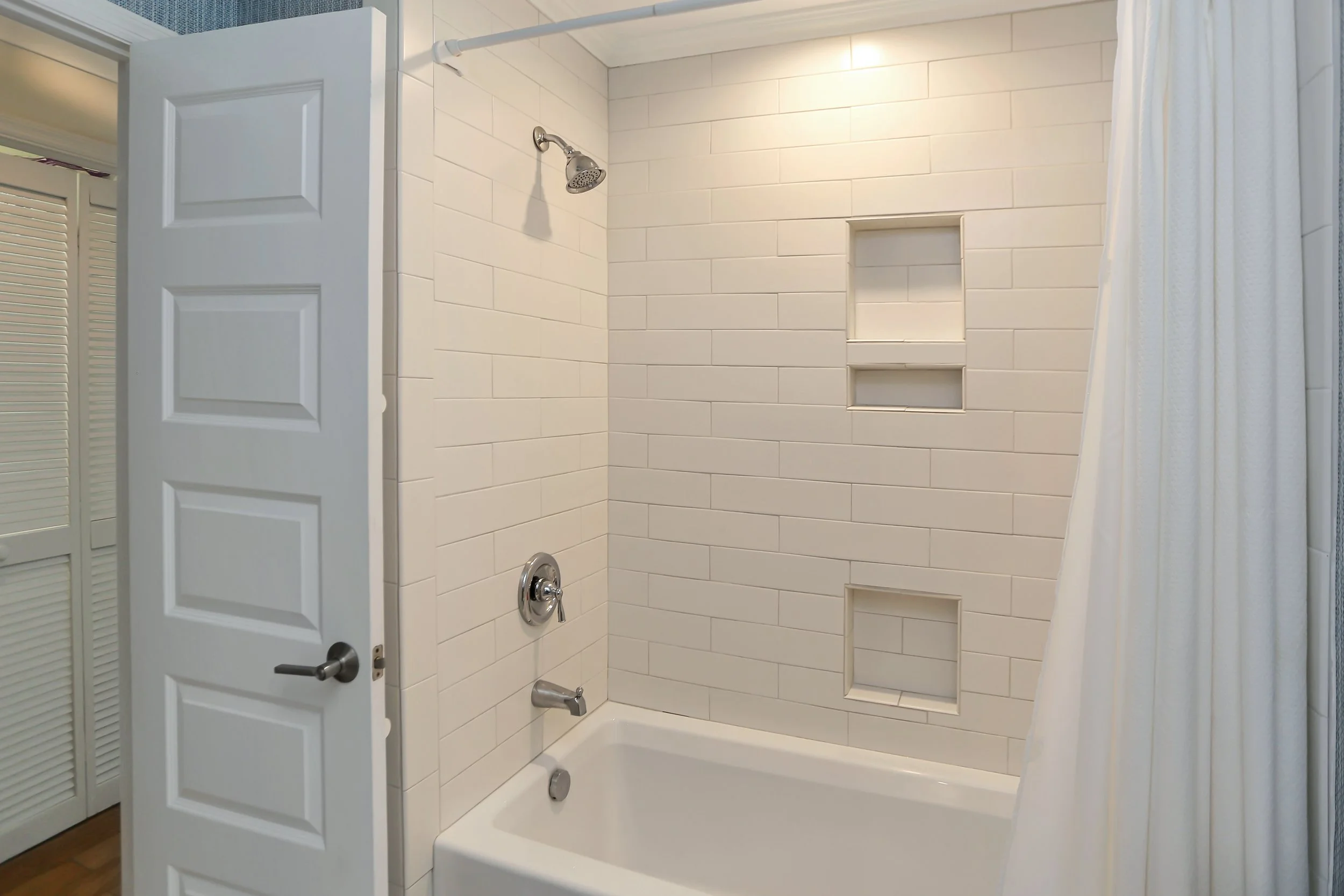 Bathroom shower with white tiled wall and built-in shelves, showerhead, and bathtub, partially covered by a white shower curtain.