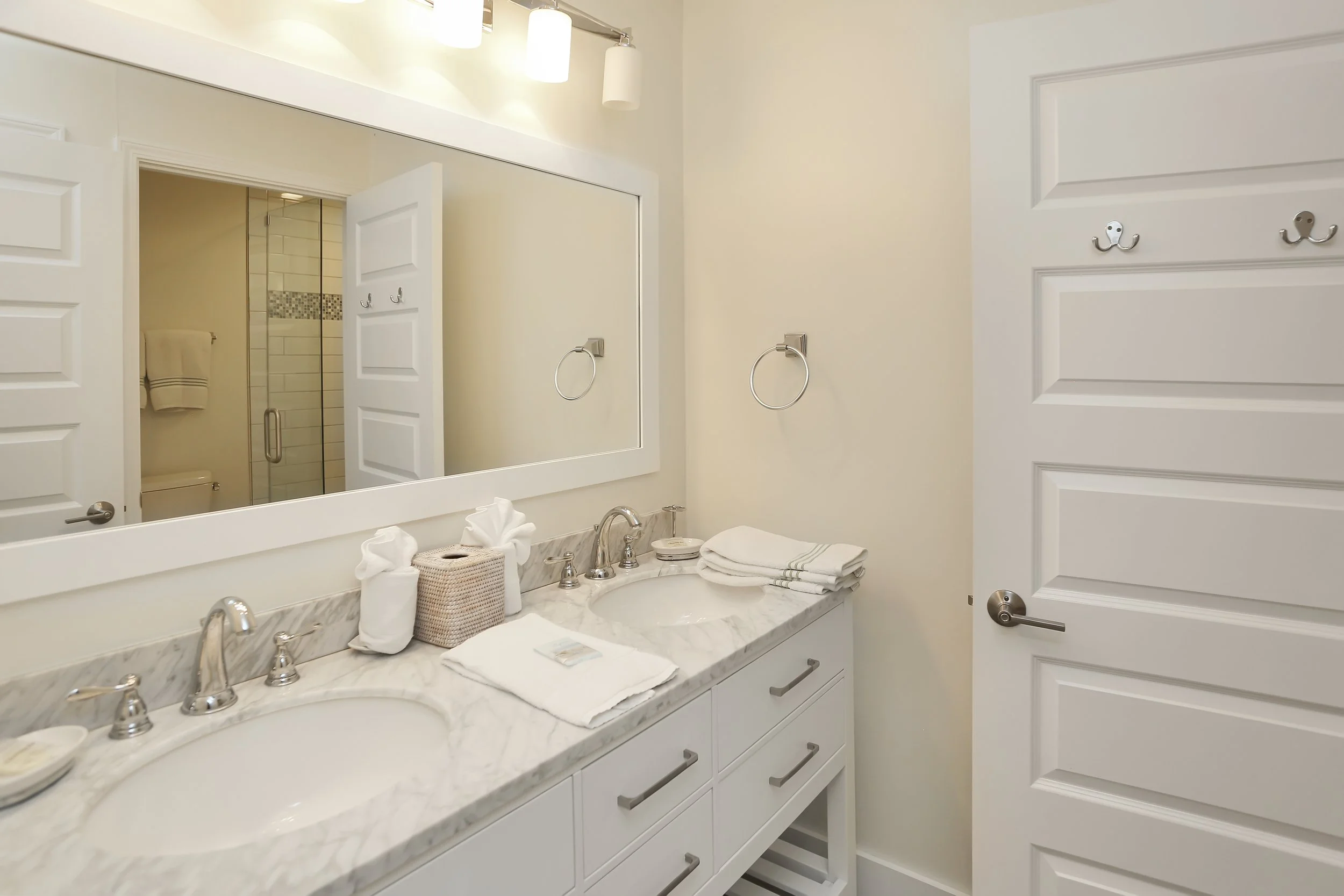 Bathroom with a double sink marble vanity, large mirror, towels, tissue box, and two towel rings, with a shower in the reflection.