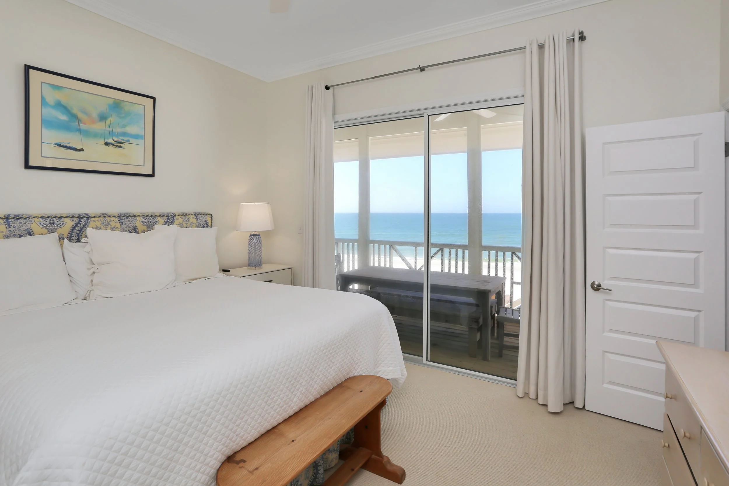 Beach view from a hotel room with a balcony, a bed with white bedding, a nightstand with a lamp, a framed picture of sailboats, and cream curtains covering the sliding glass door.