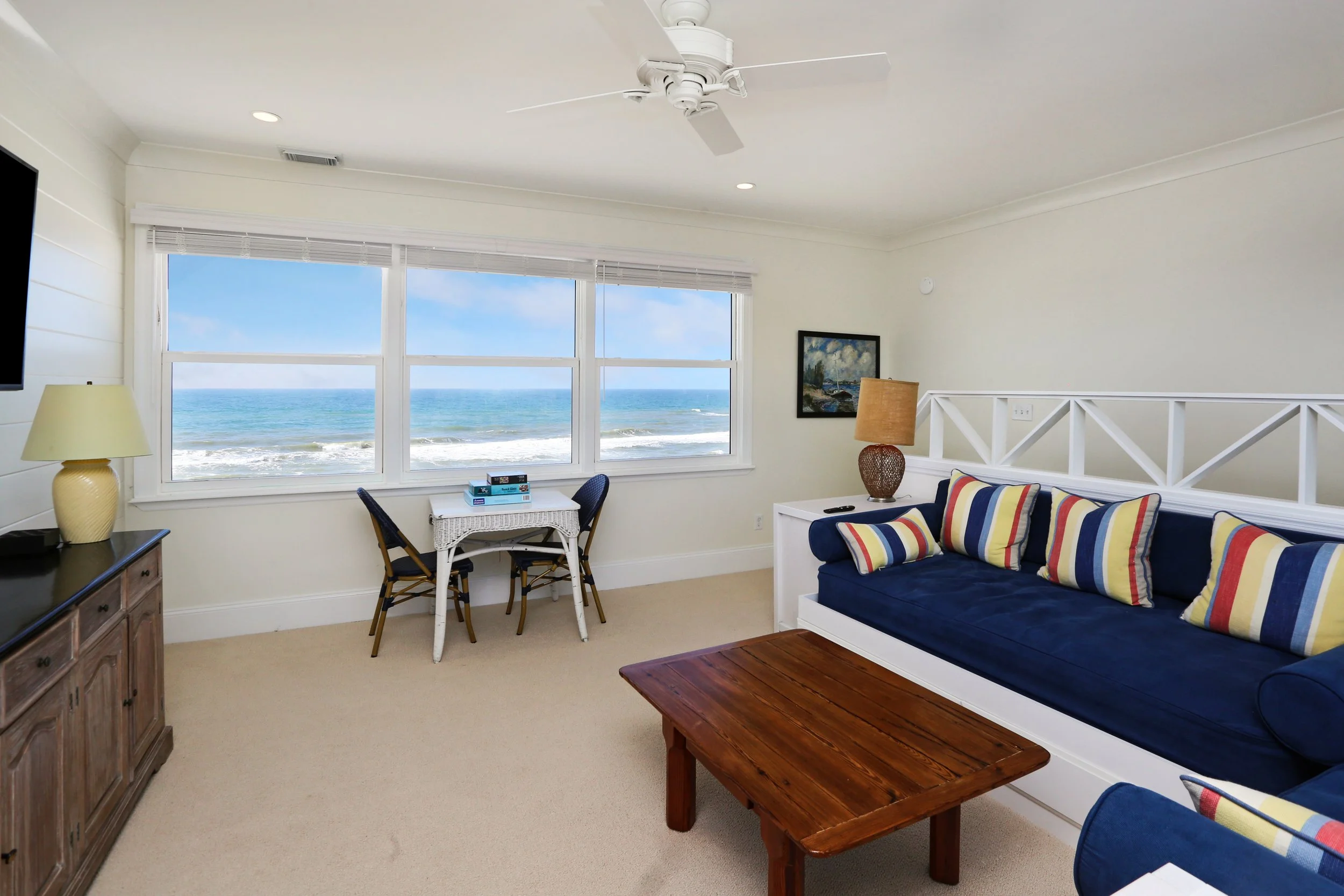 Living room with large window overlooking the ocean, navy sofa with striped pillows, wooden coffee table, side table with lamp, and small table with chairs near the window.