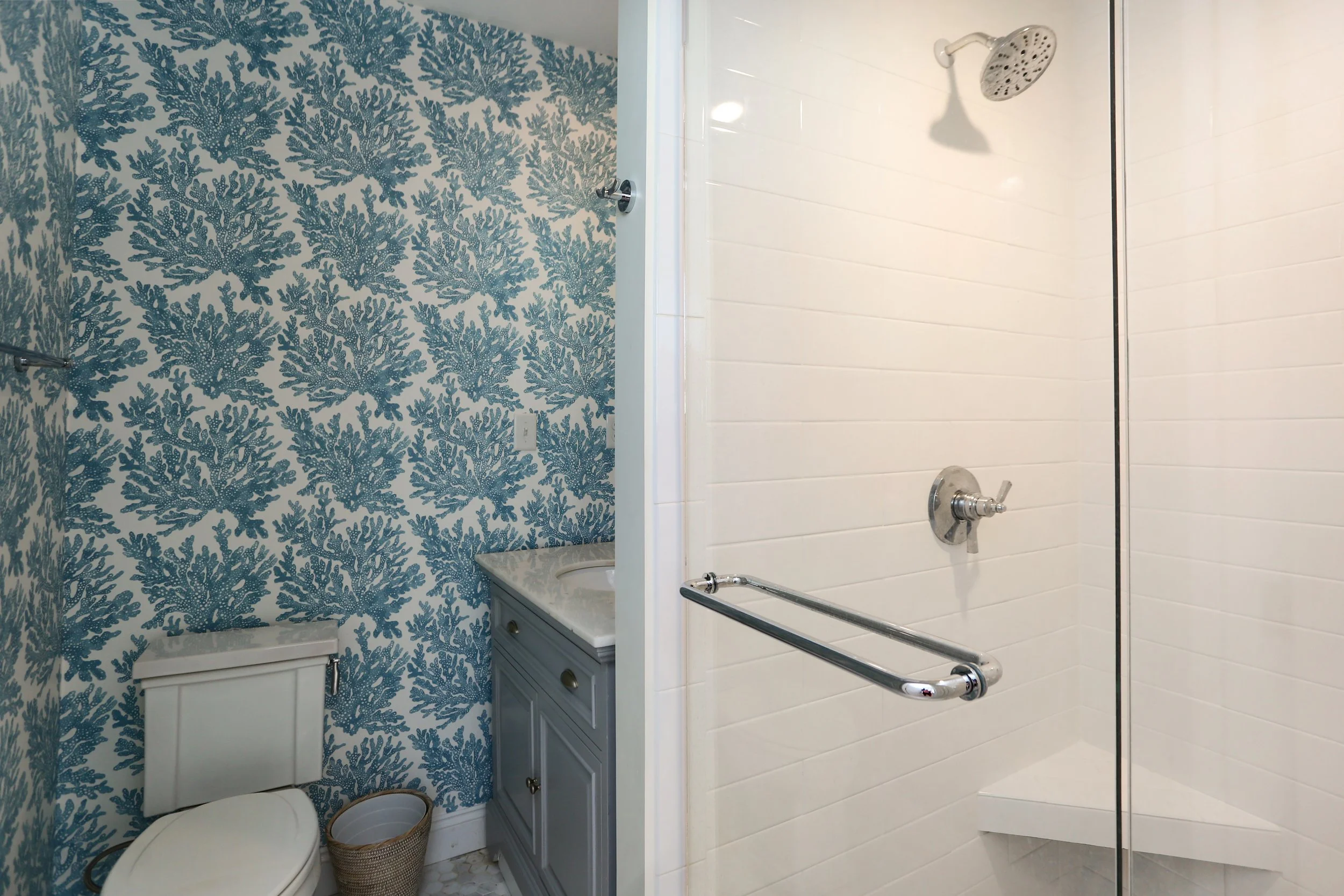 Bathroom with blue coral-patterned wallpaper, toilet, small vanity with sink, and walk-in shower with white tiled walls and chrome fixtures.