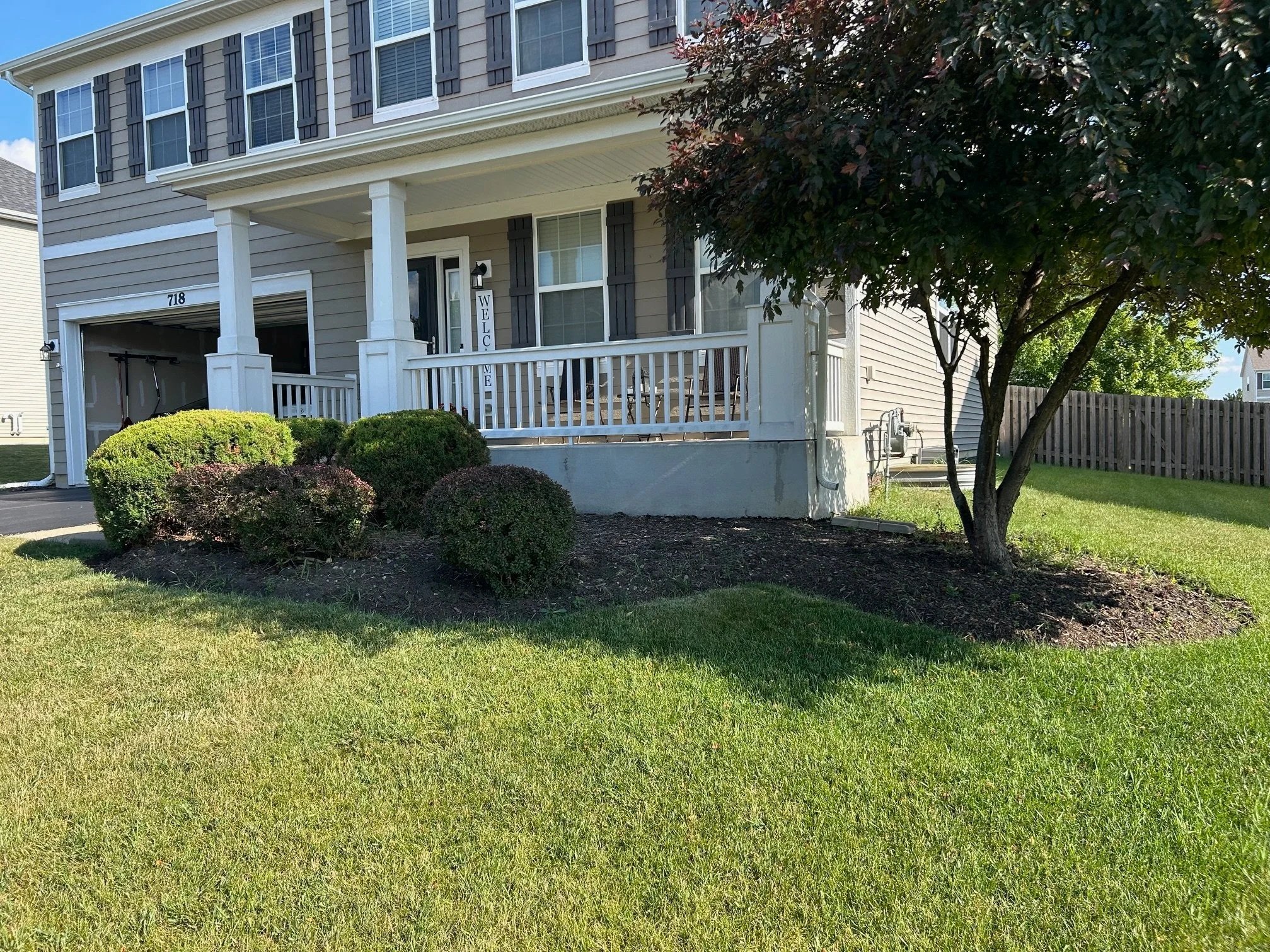 Front yard of a house with green grass, landscaped bushes, a tree, and a porch with white railings.