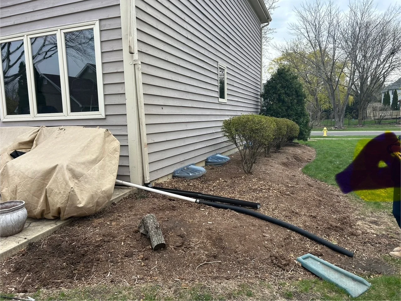 Side yard with bushes, a house with gray siding, a small window, and part of a yard and street with a fire hydrant in the background. There are gardening tools and materials, including a covered object, pipes, and a log on the ground.
