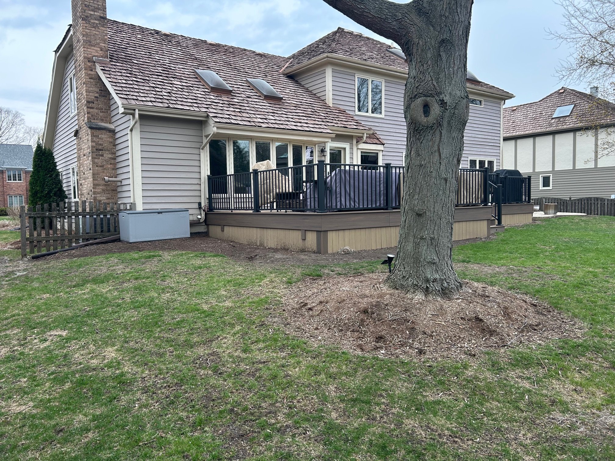 Backyard of a house with a large tree, a deck with black railing, and a grassy lawn.