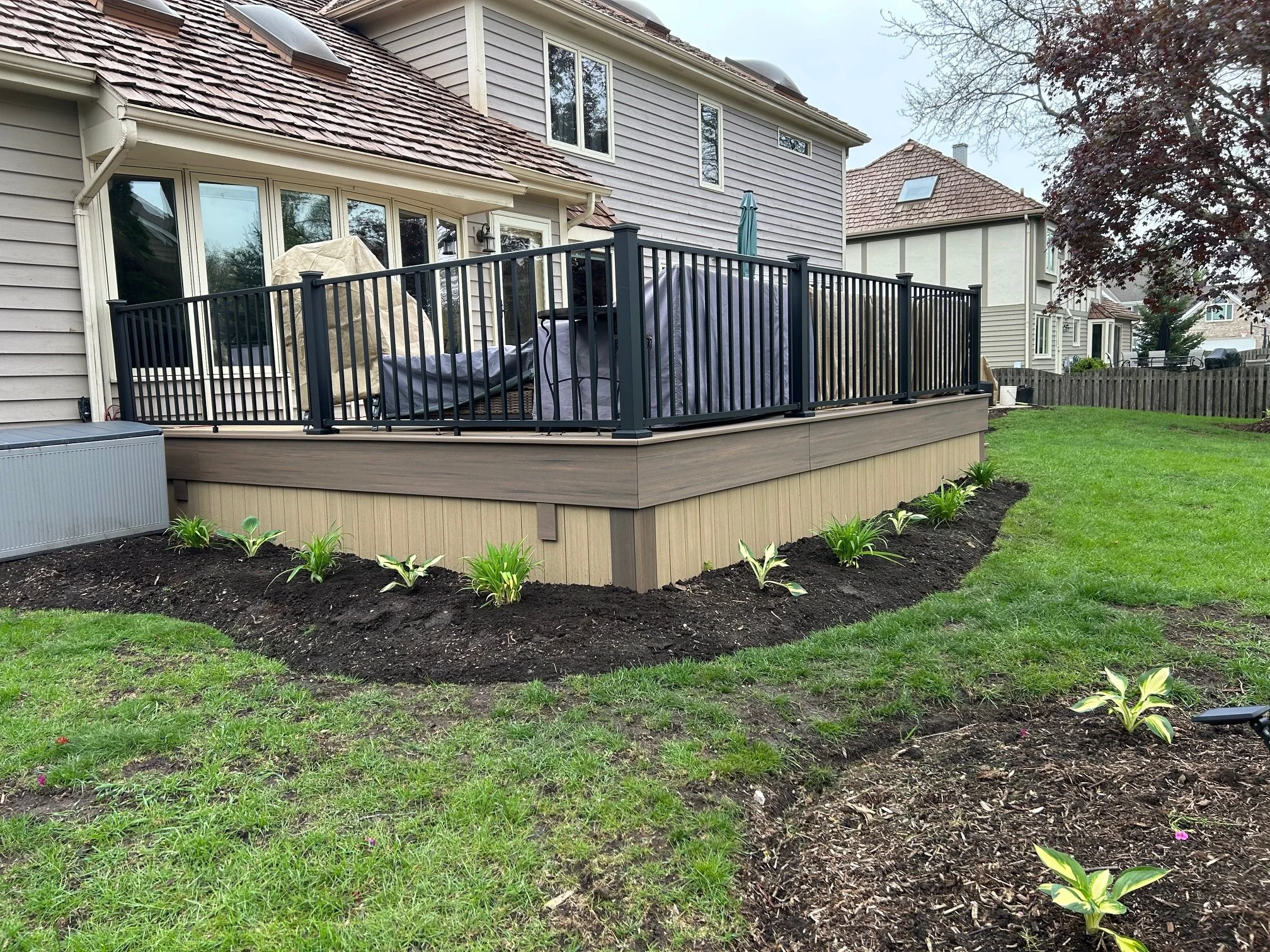 A backyard patio with a black metal railing, outdoor furniture, and a covered grill, surrounded by a garden with newly planted flowers and green grass.