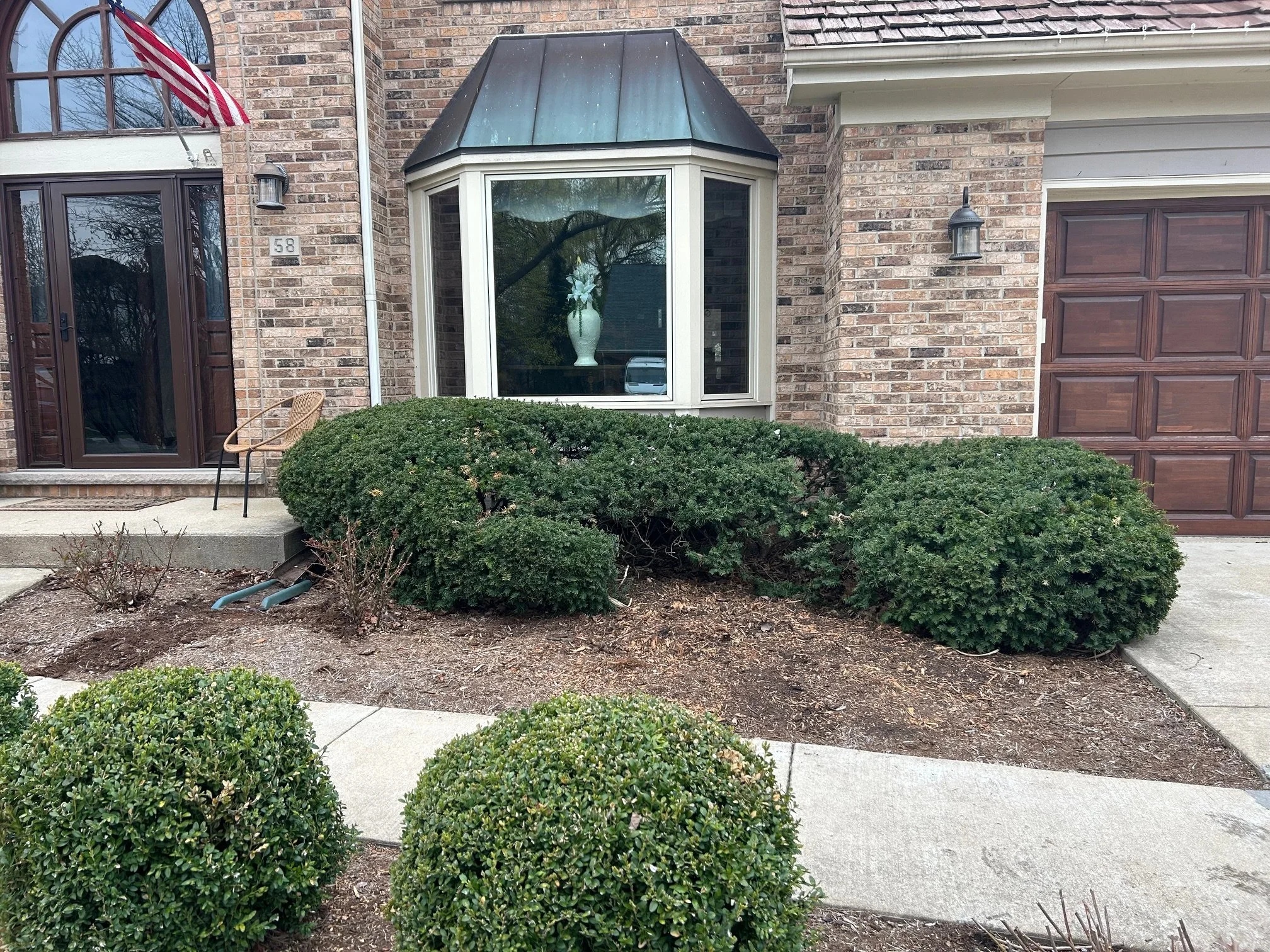 The front yard of a brick house with a large bay window, decorative bushes, and a concrete walkway leading to the entrance.