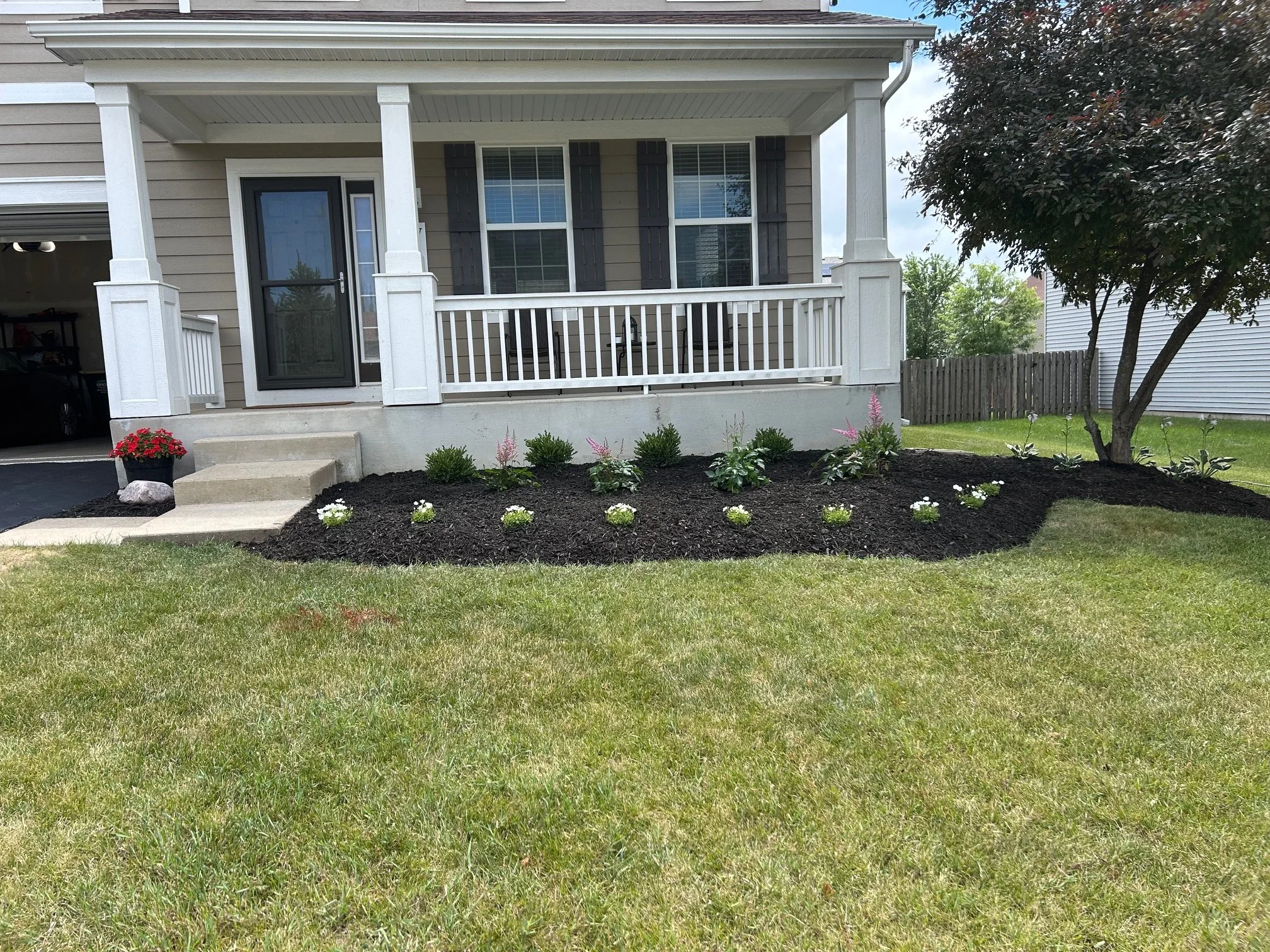 Front porch of a house with a small set of stairs, a white railing, and a porch with a charcoal gray door. The yard includes freshly planted flower beds with small white flowers, pink flowers, green shrubs, a large tree to the right, and a lush green