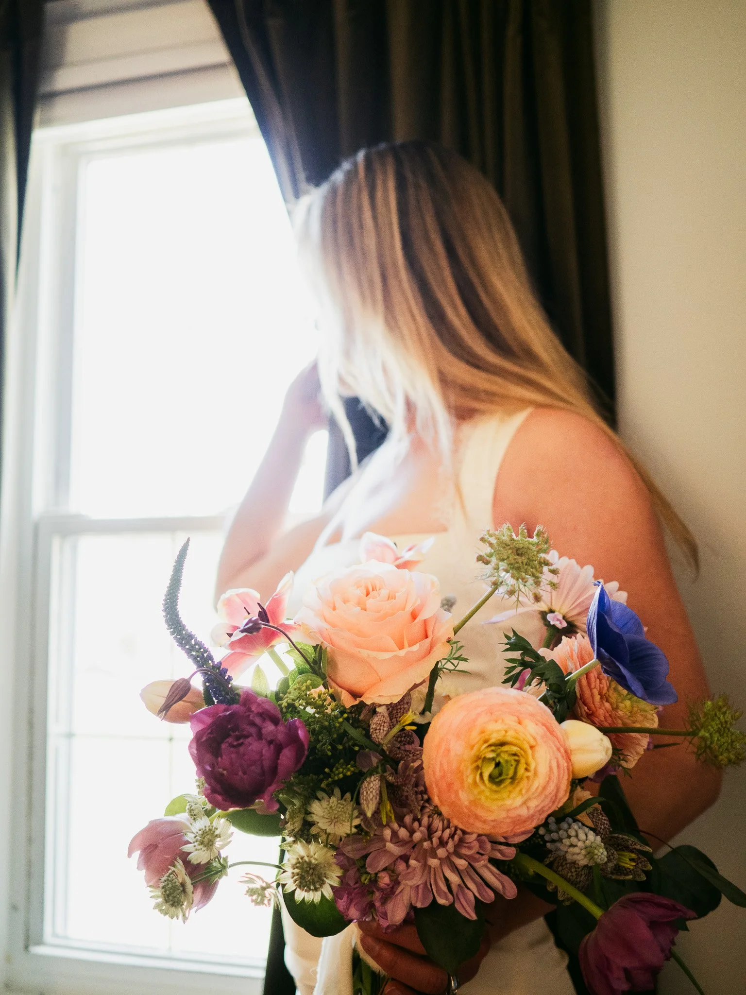 Person holding a large bouquet of colorful flowers, including roses, peonies, and other flowers, near a window with dark curtains.