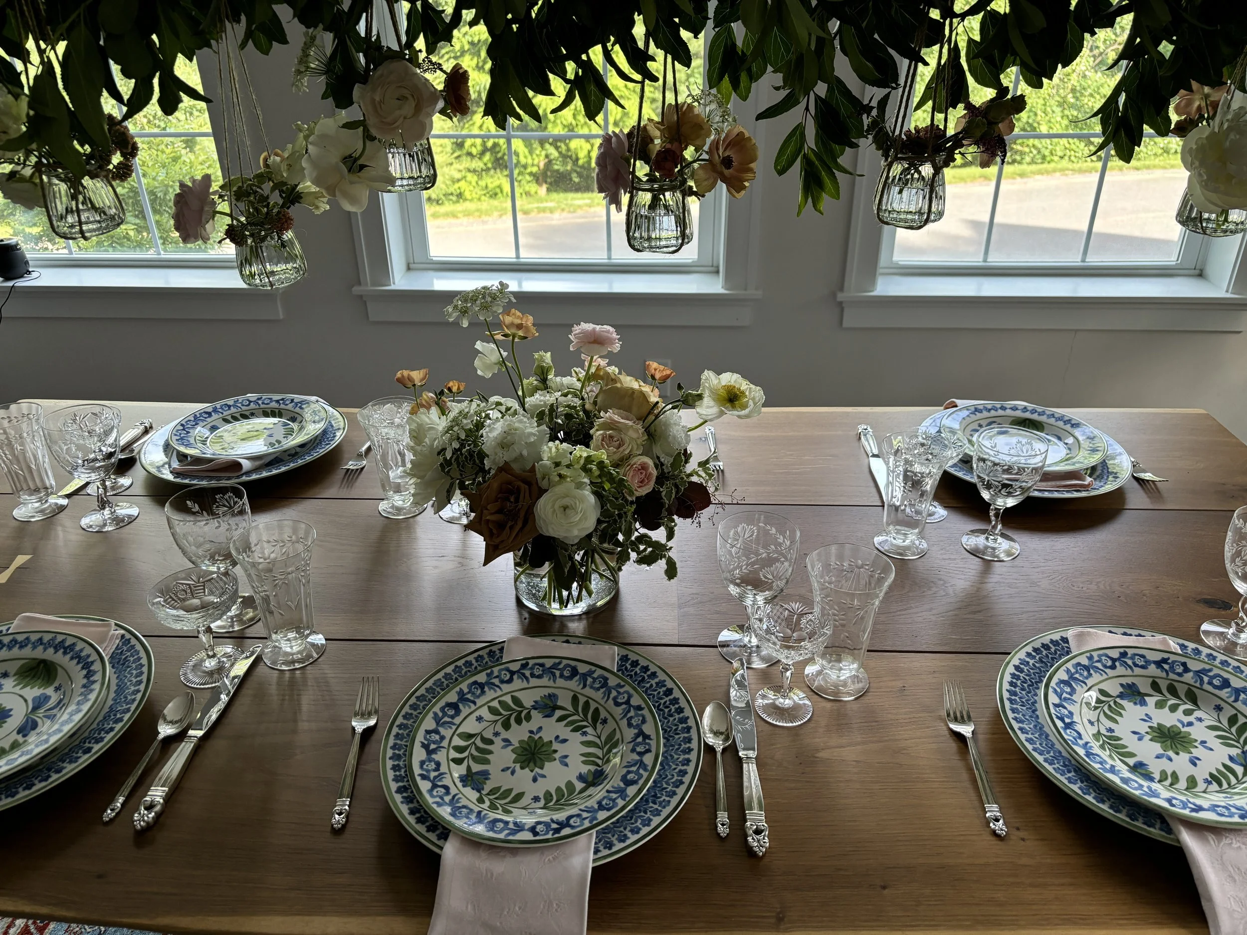 A dining table set for a meal with floral-patterned plates, silverware, and glasses, featuring a centerpiece of assorted flowers in a glass vase, with large windows and hanging floral arrangements overhead.