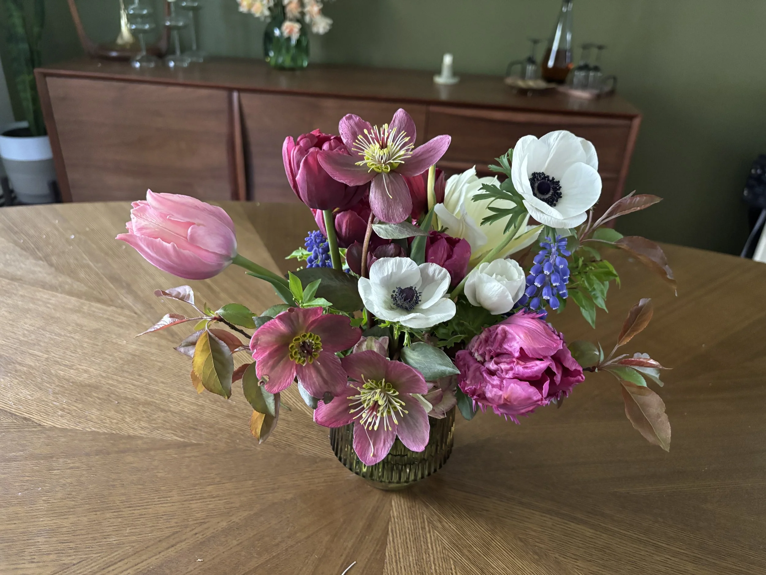 A bouquet of colorful flowers in a glass vase on a wooden table in a room with a green wall and a wooden sideboard in the background.