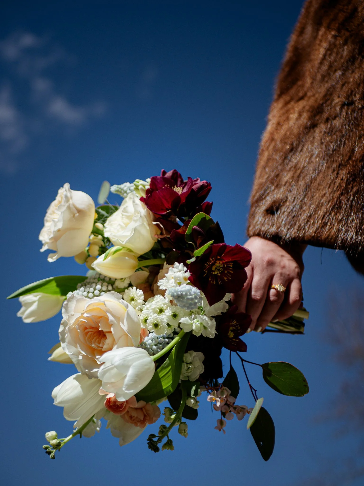 Person holding a wedding bouquet of various flowers against a bright blue sky.