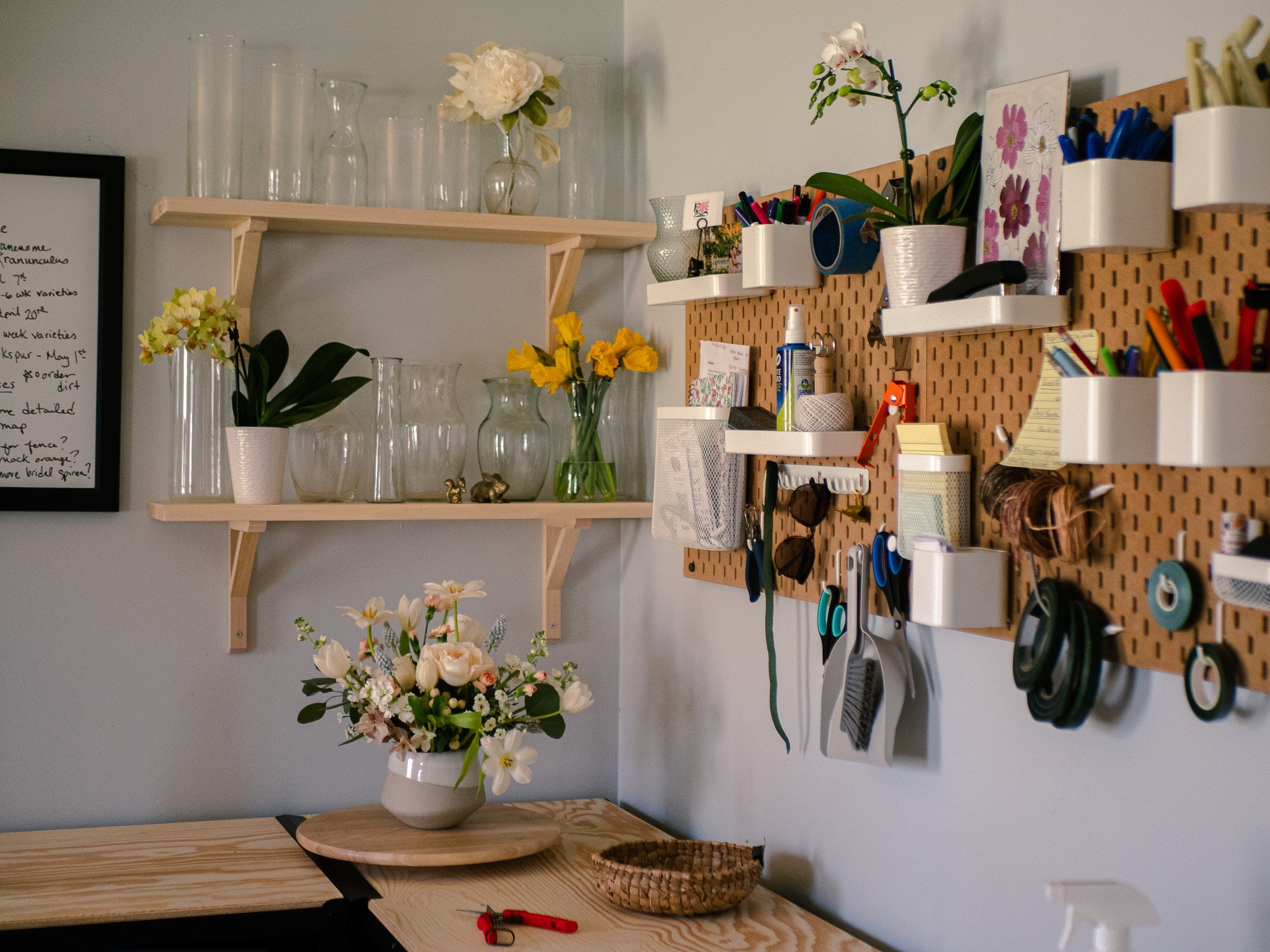 A home workspace with floral arrangements on a light wood desk and white shelves, organized with pens, scissors, and office supplies.
