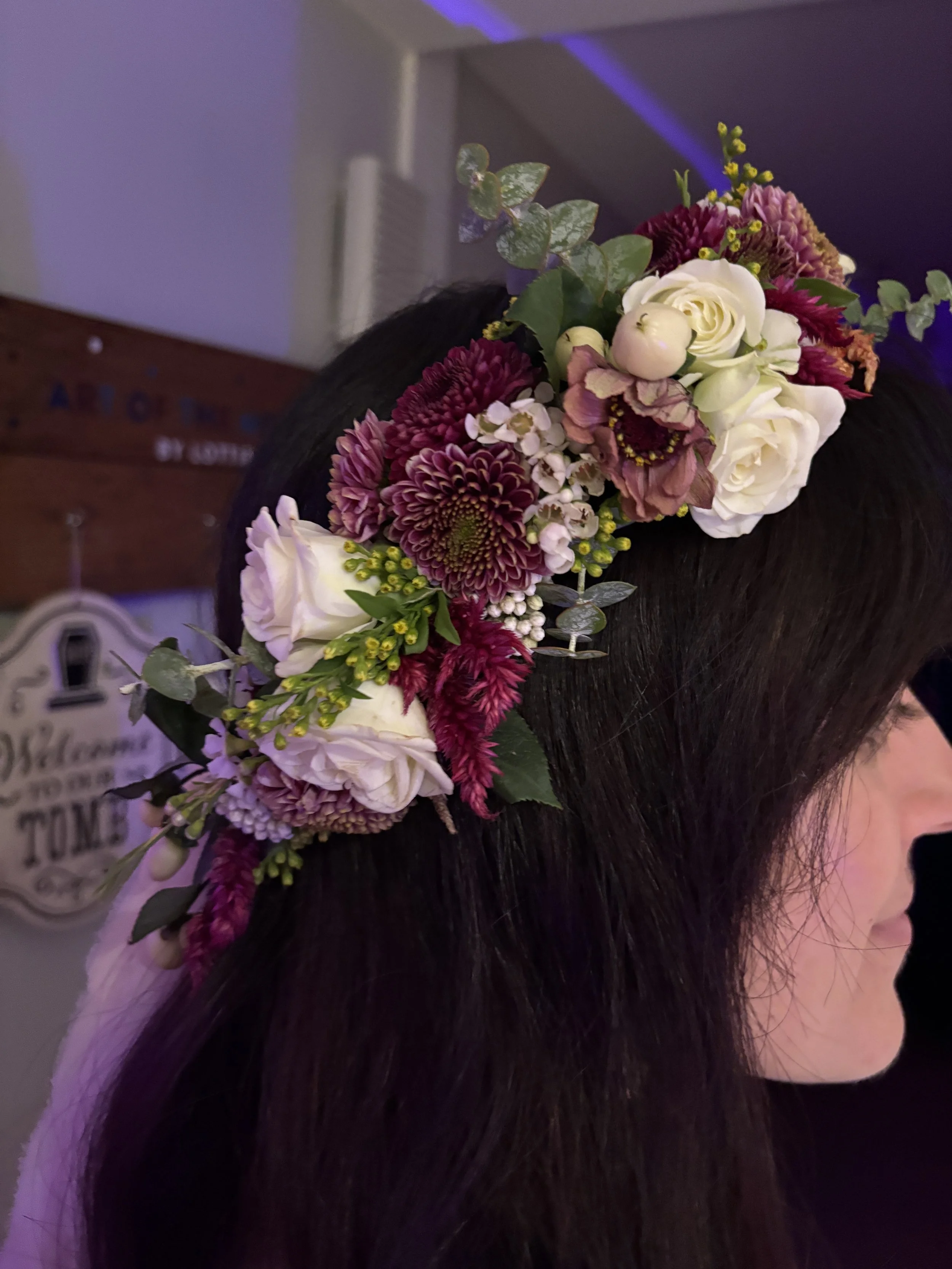 A floral crown made of white roses, deep red dahlias, and purple and pink flowers with green foliage, worn on a person's head with dark hair.