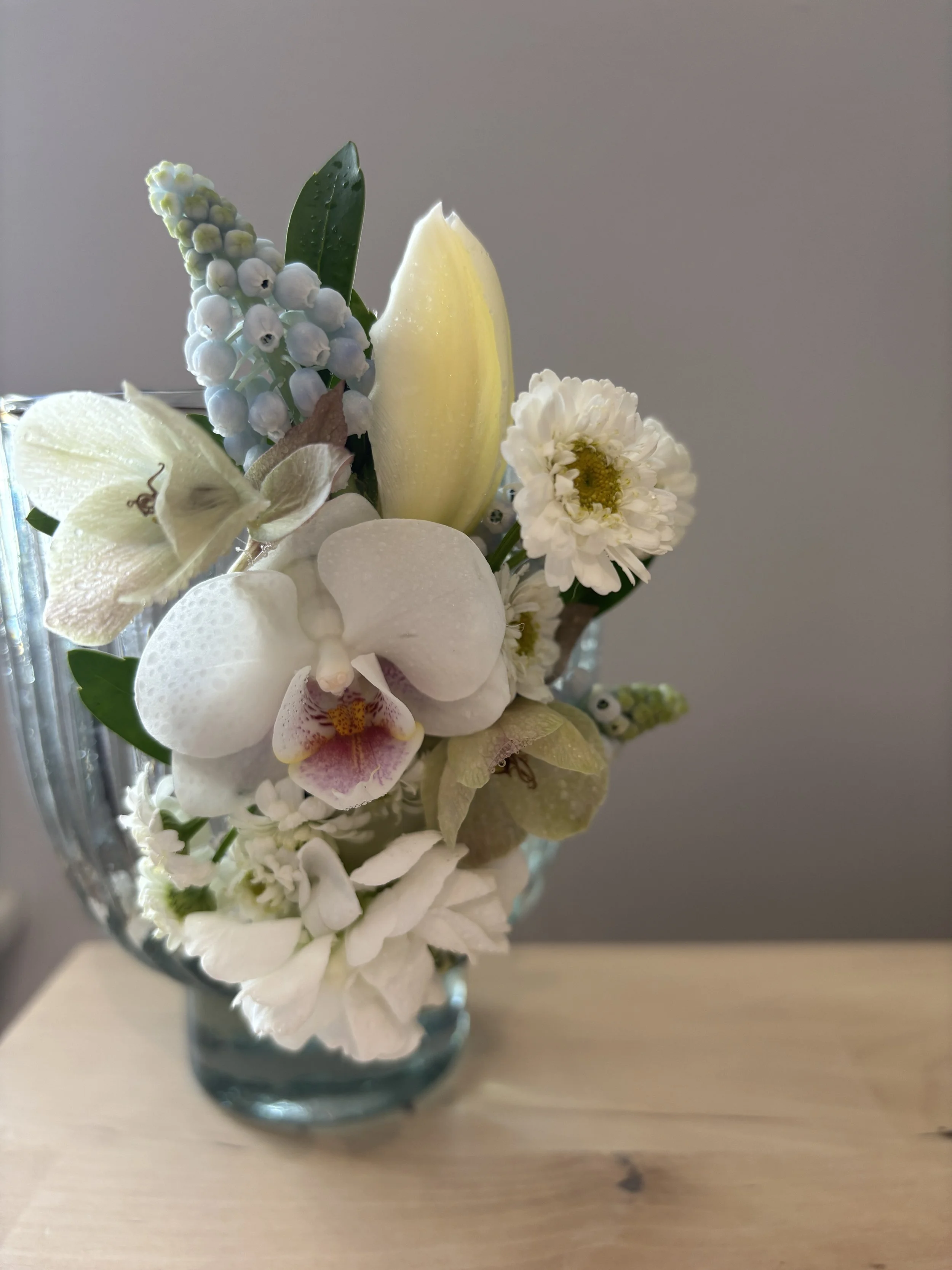 A button boutonniere in a glass vase containing white orchids, a white flower with a yellow center, green leaves, and a white flower with purple markings.