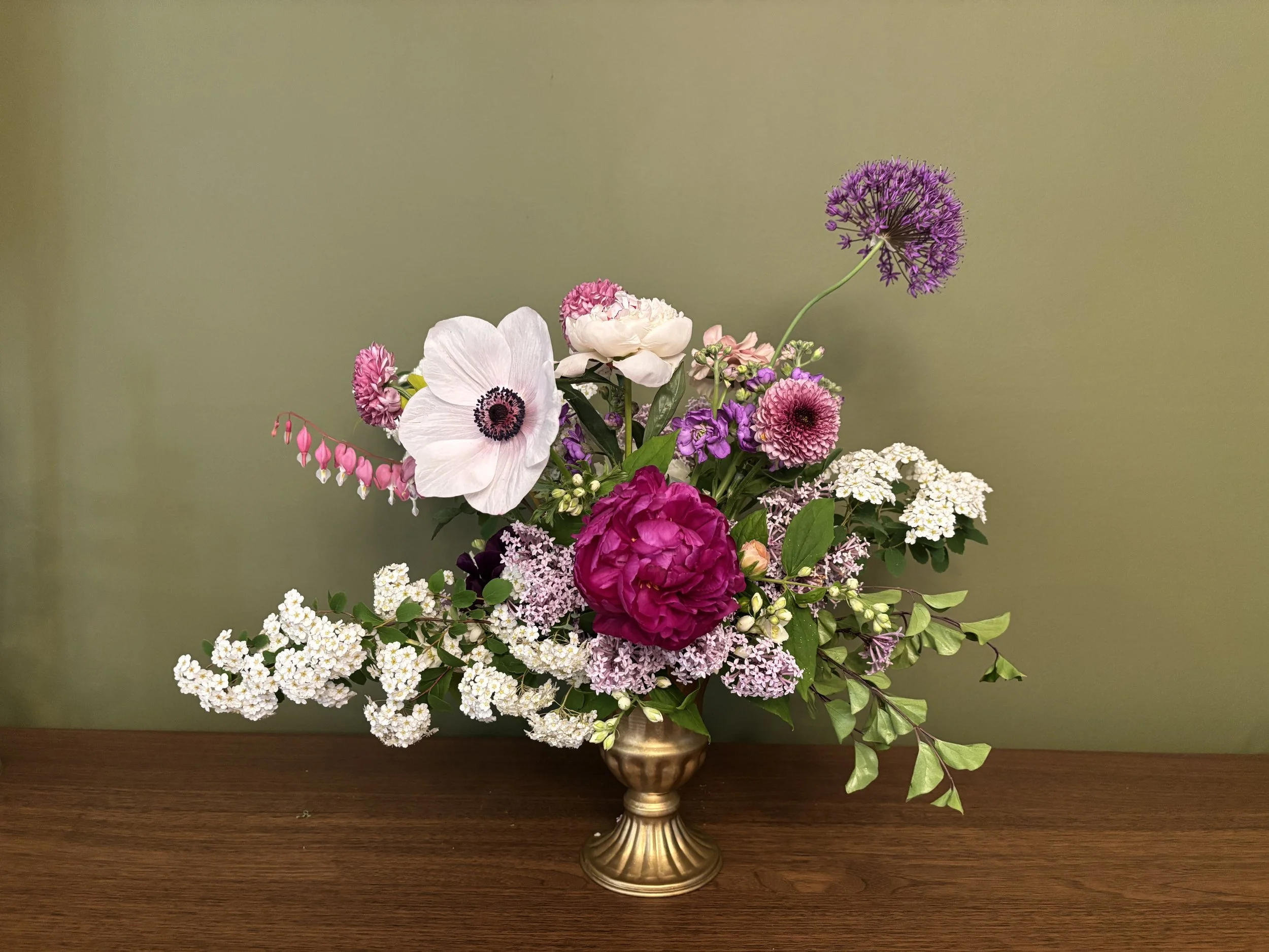 A floral arrangement in a gold decorative vase featuring pink, white, purple, and green flowers against a plain background, resting on a wooden surface.