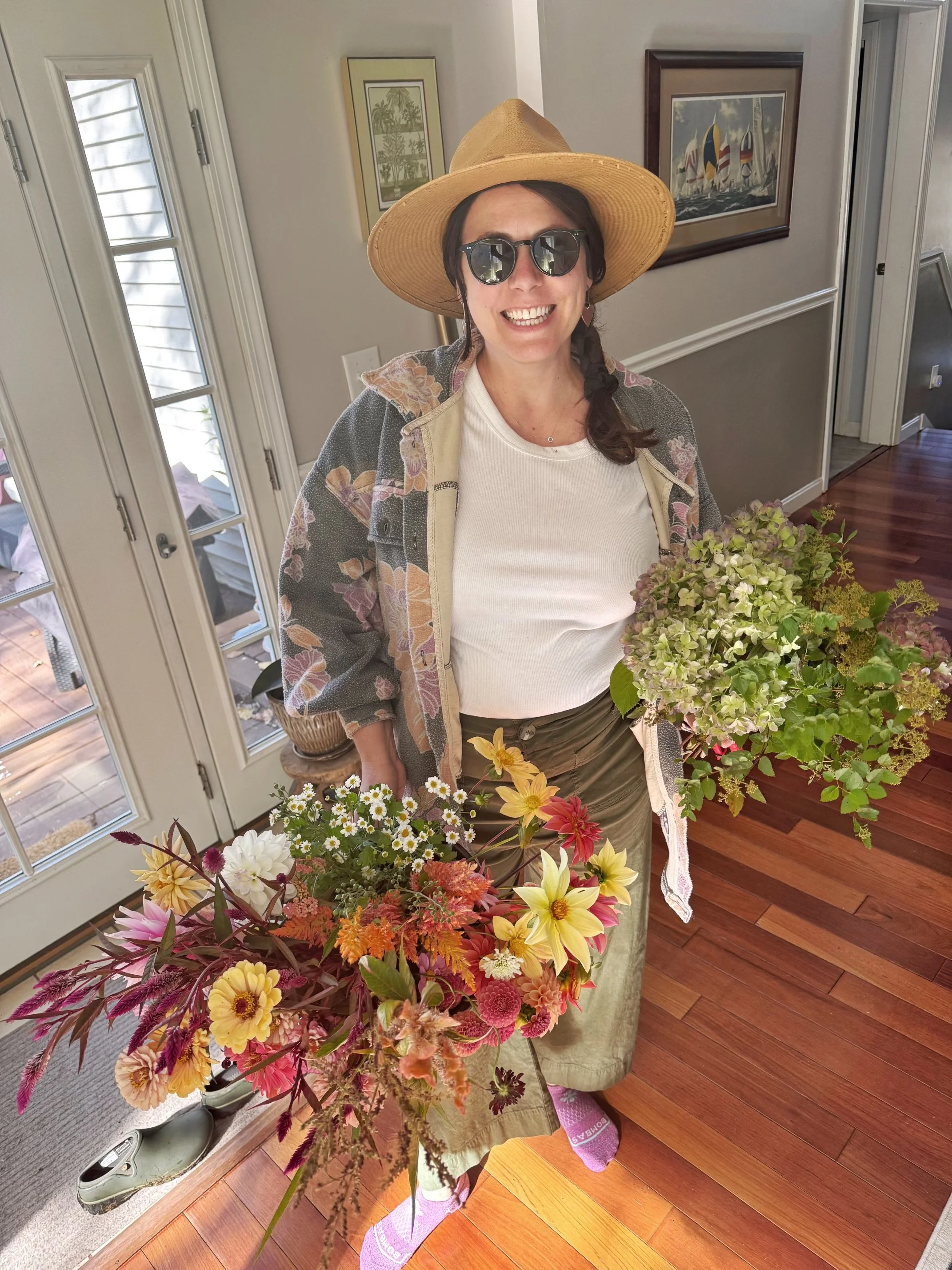 A woman smiling, wearing sunglasses and a large straw hat, holding a buckets of colorful freshly harvested flowers inside a house.