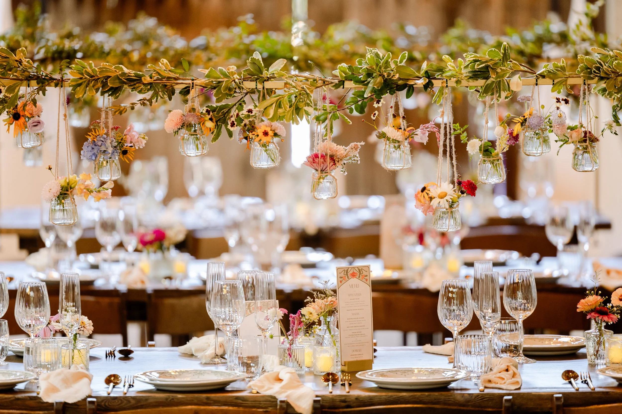 A banquet table decorated with hanging glass jars filled with colorful flowers and greenery, set for a formal event with plates, glasses, and table settings.