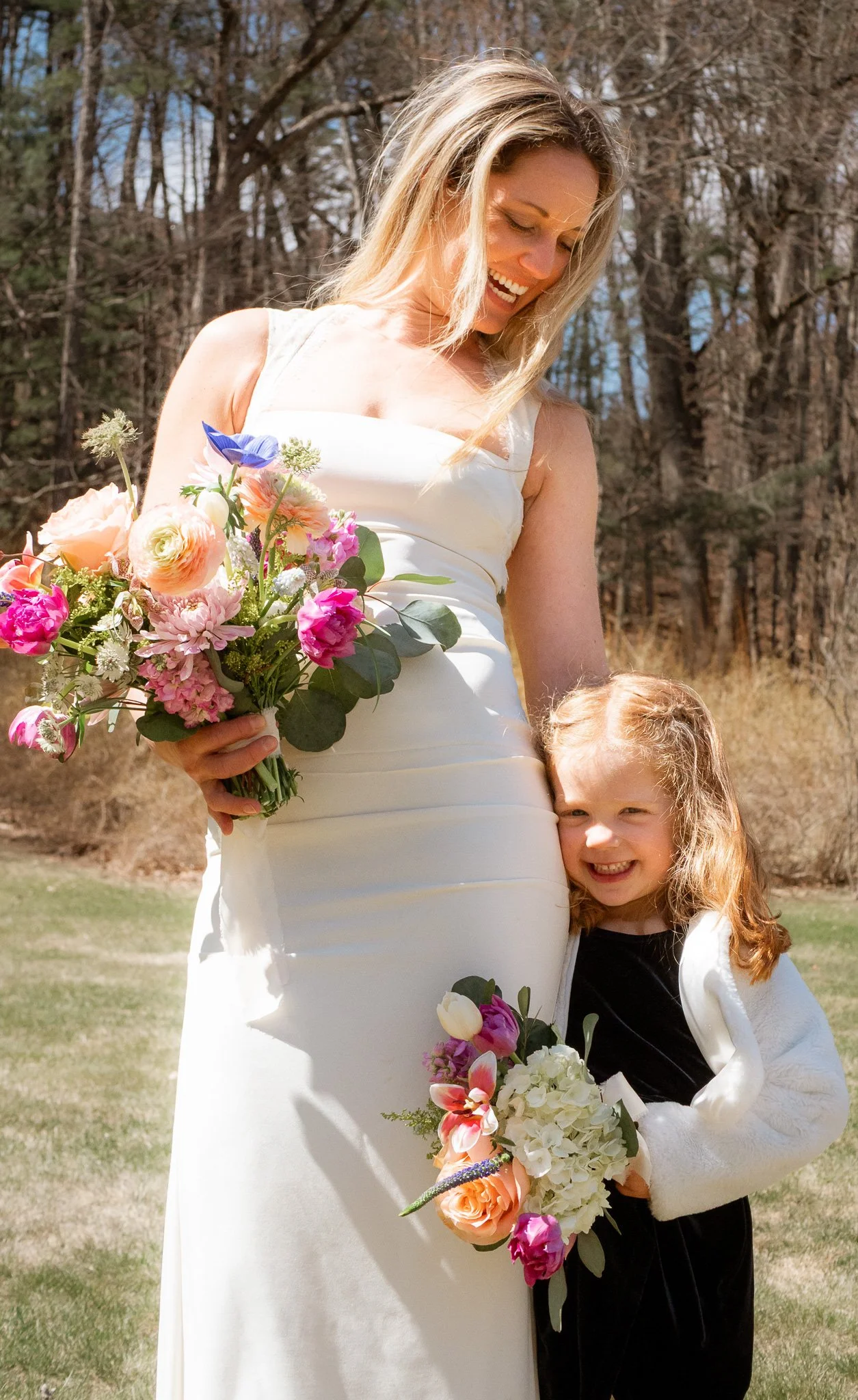 A woman in a white dress holding a colorful bouquet of flowers, smiling, standing outdoors with a young girl with curly red hair holding a bouquet, both smiling, in a park or garden area with trees in the background.
